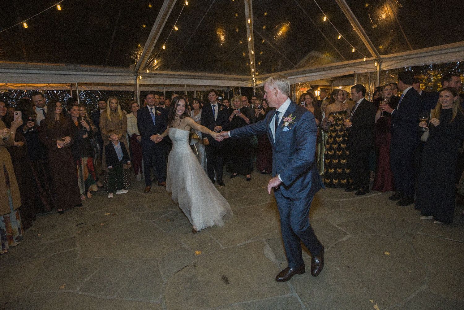 A bride in a white gown and a groom in a navy suit share a first dance under a tent as wedding guests watch.