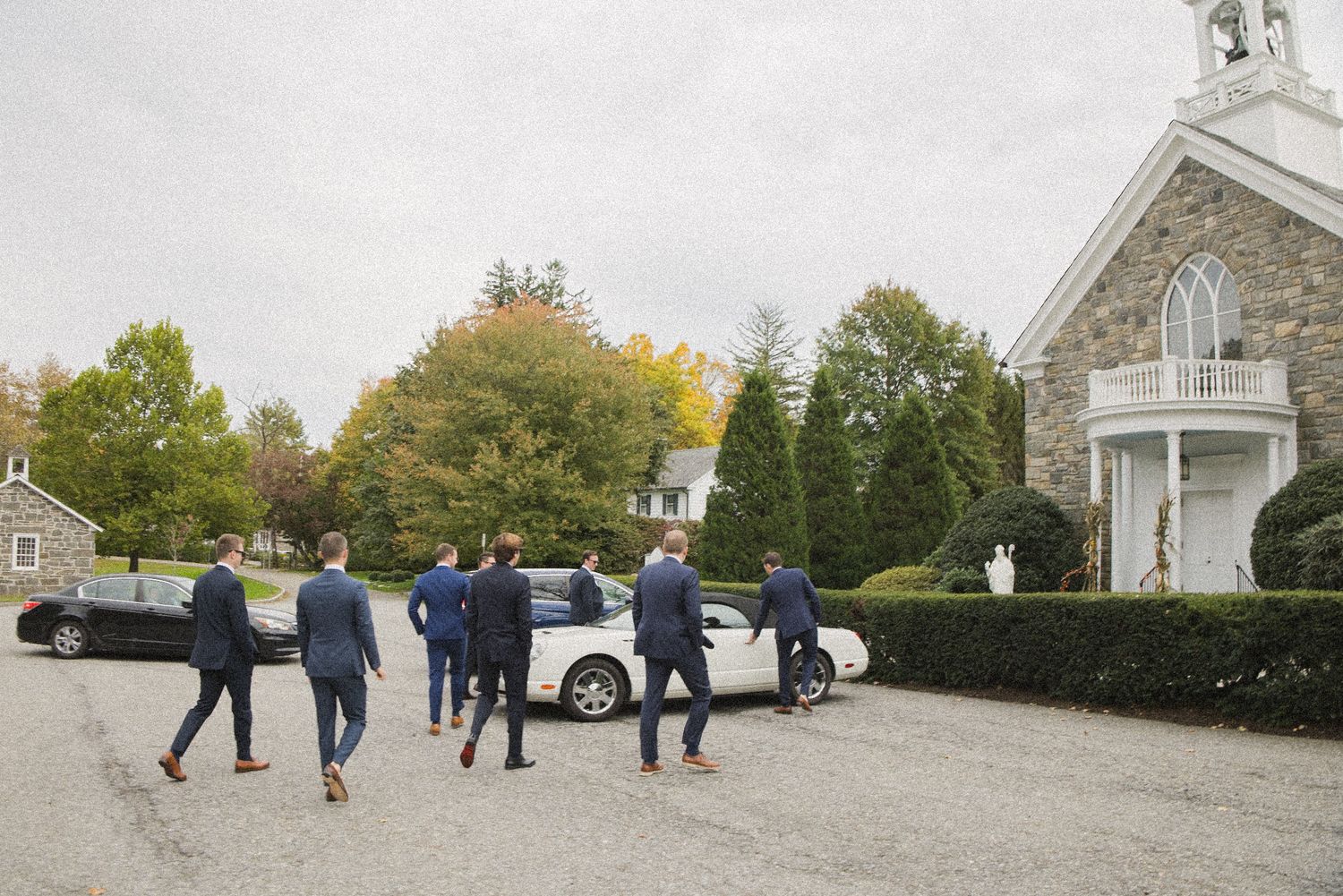 Groomsmen in navy suits walk across a gravel lot toward a white car beside a historic stone church surrounded by colorful autumn trees.