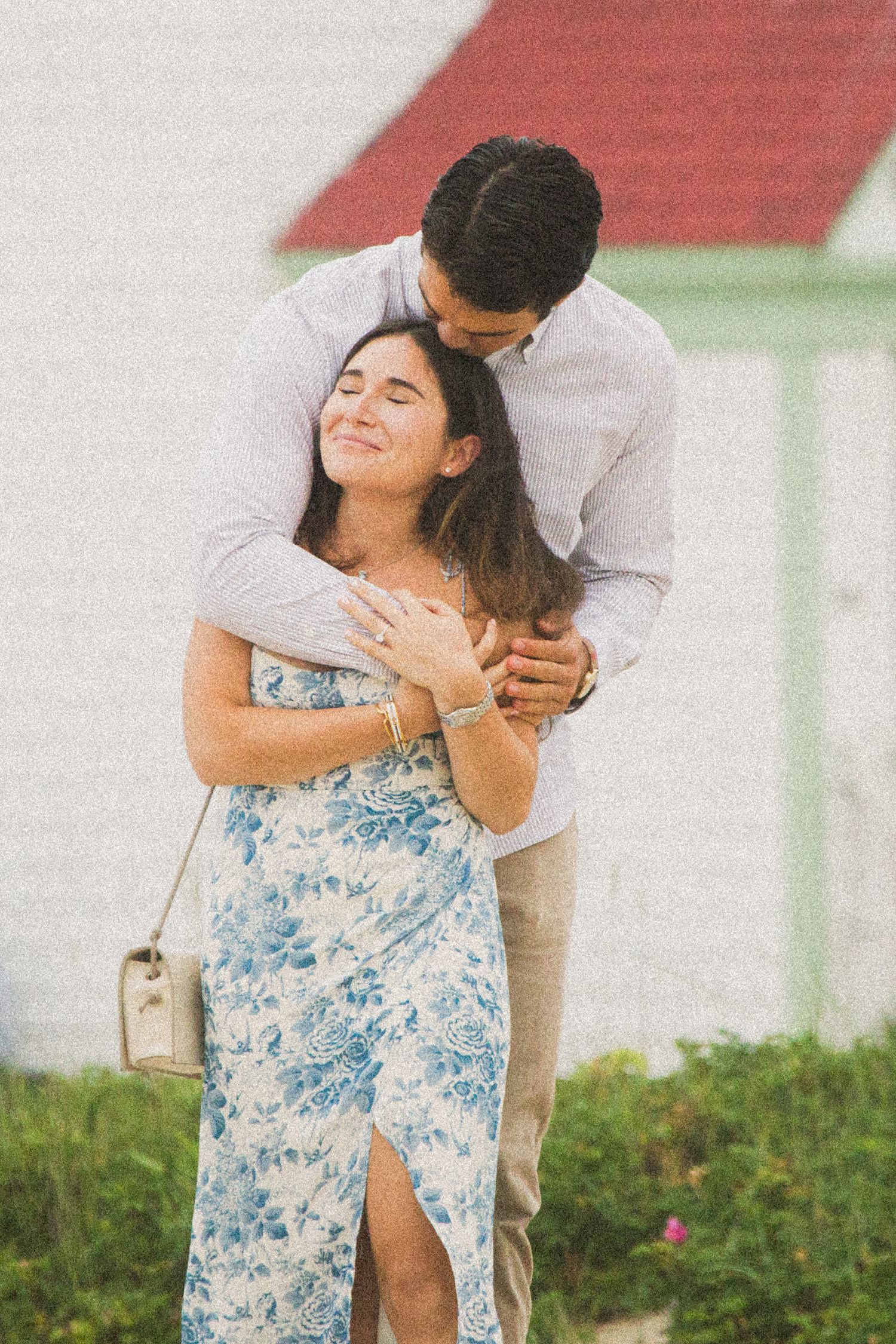 Couple embracing outdoors, woman in blue floral dress smiling with eyes closed as man hugs her from behind.