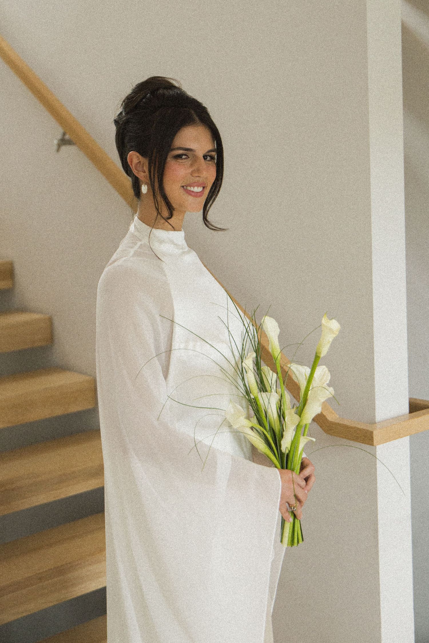 Elegant bride in white high-neck gown holding calla lily bouquet, standing by wooden staircase, smiling softly.