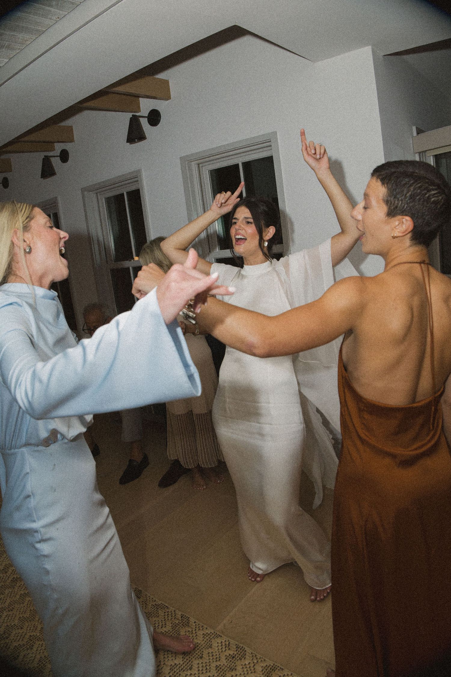 Women in elegant attire dancing joyfully at a wedding reception, with bride in white dress raising arms.