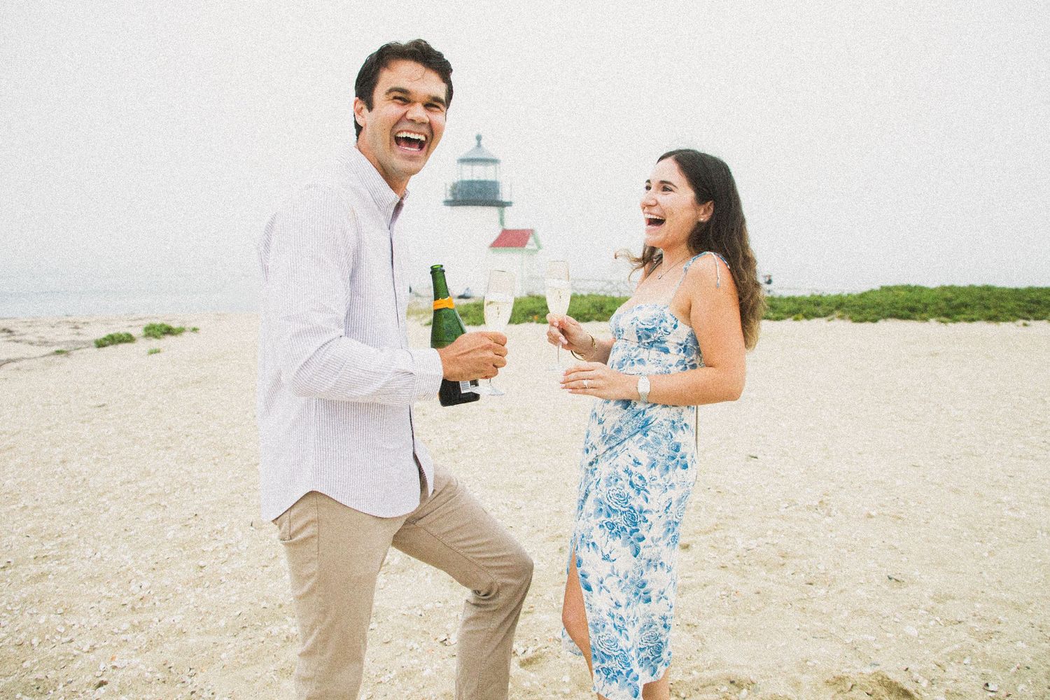 A couple celebrates joyfully on a beach with champagne and a lighthouse visible in the background.