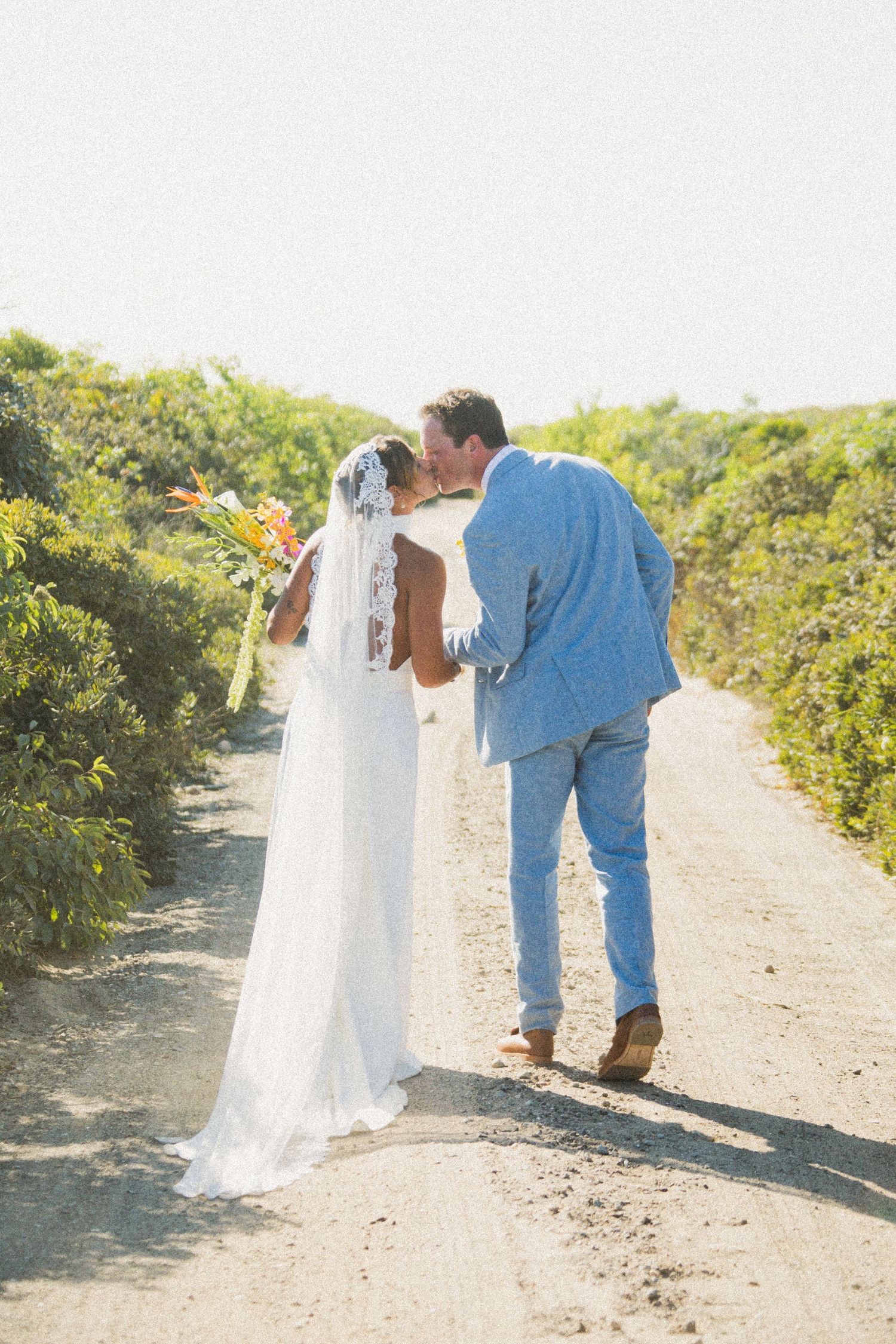 Wedding couple walks together on a sunlit dirt path with green shrubs lining both sides.