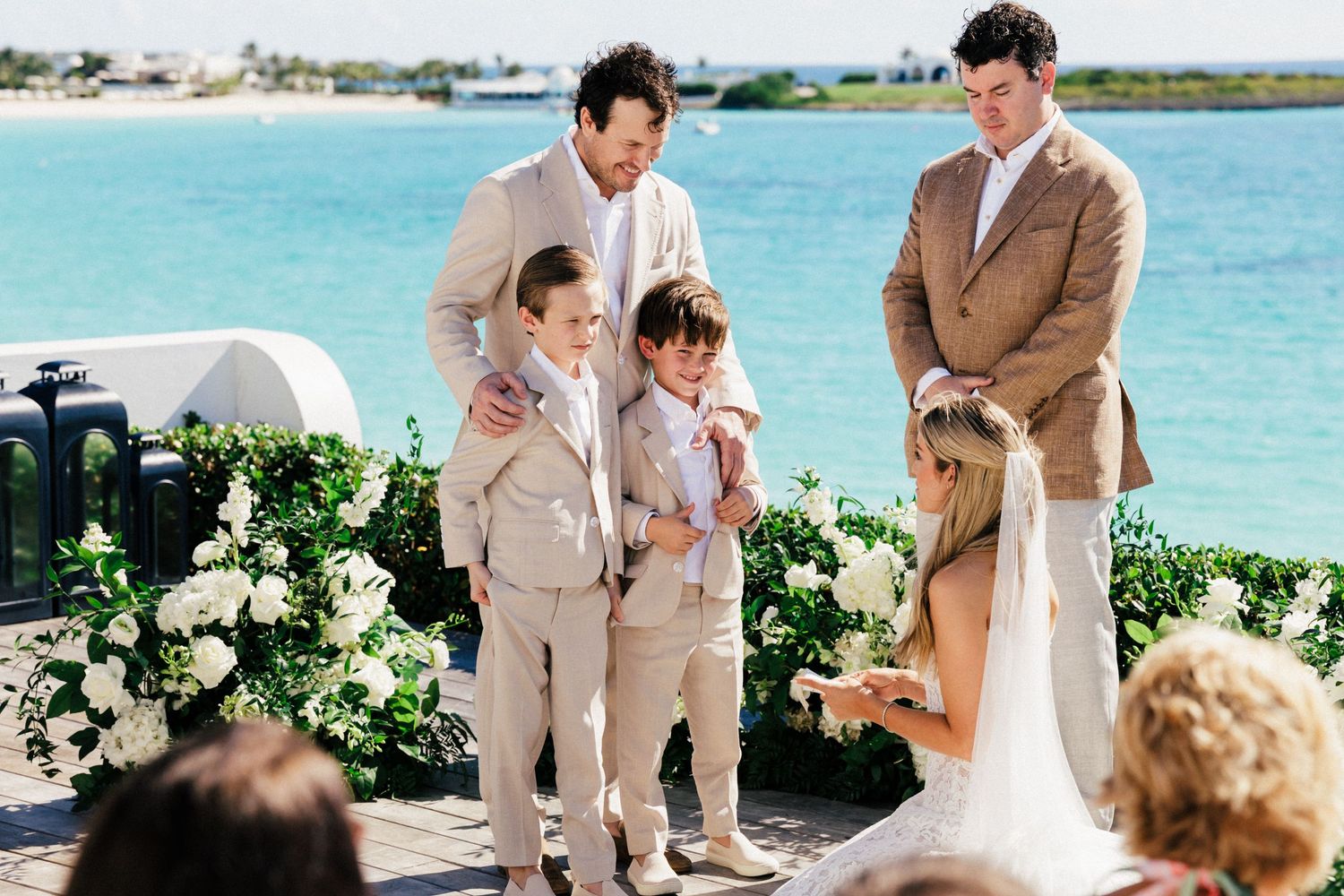 Intimate beach wedding ceremony with attendees in beige suits overlooking crystal blue waters.