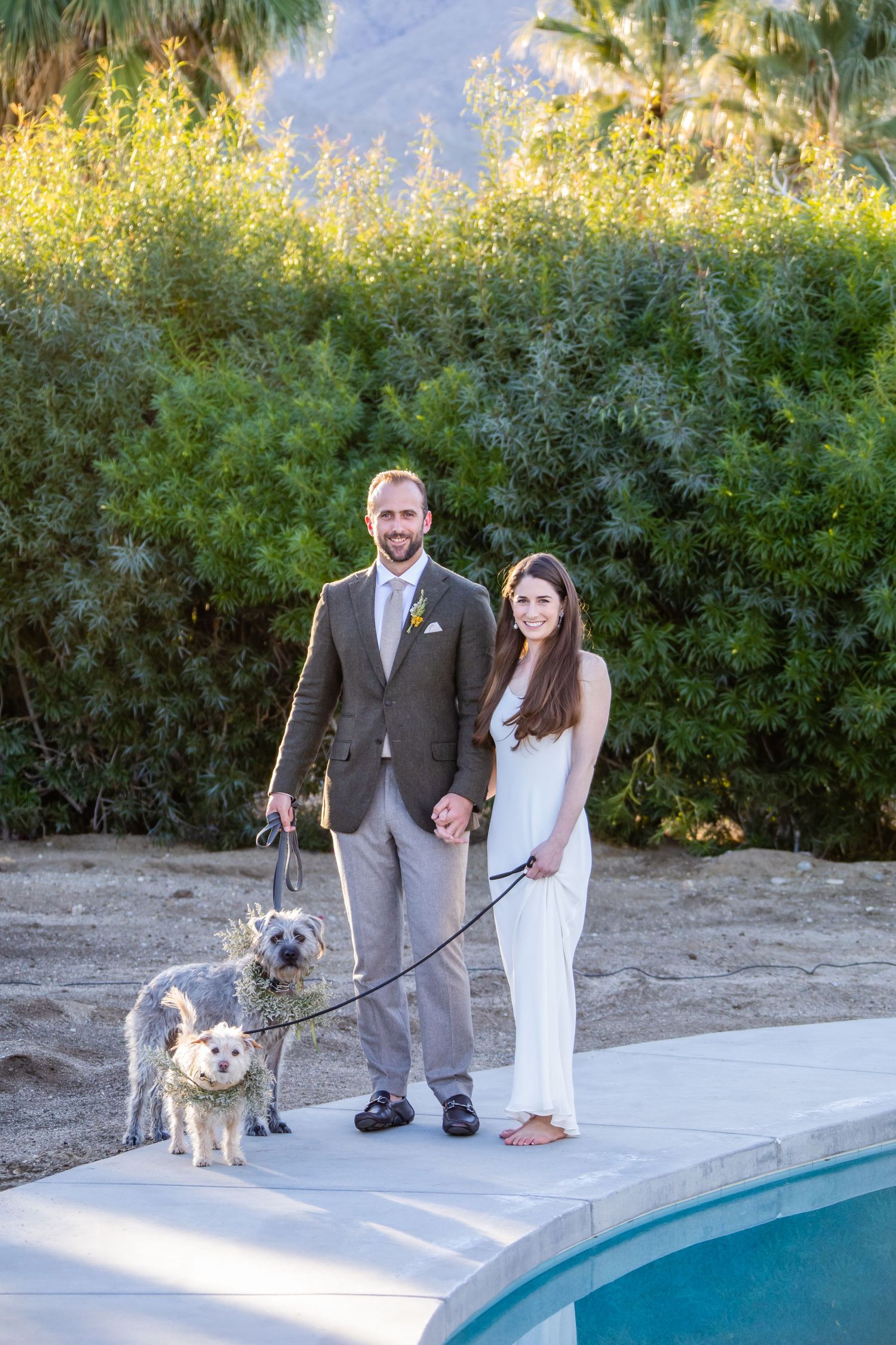 A couple poses with their two small dogs by a swimming pool with mountains in the background.