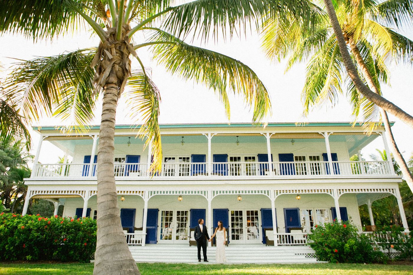 Colonial style white mansion with wraparound porch and palm trees in tropical setting.