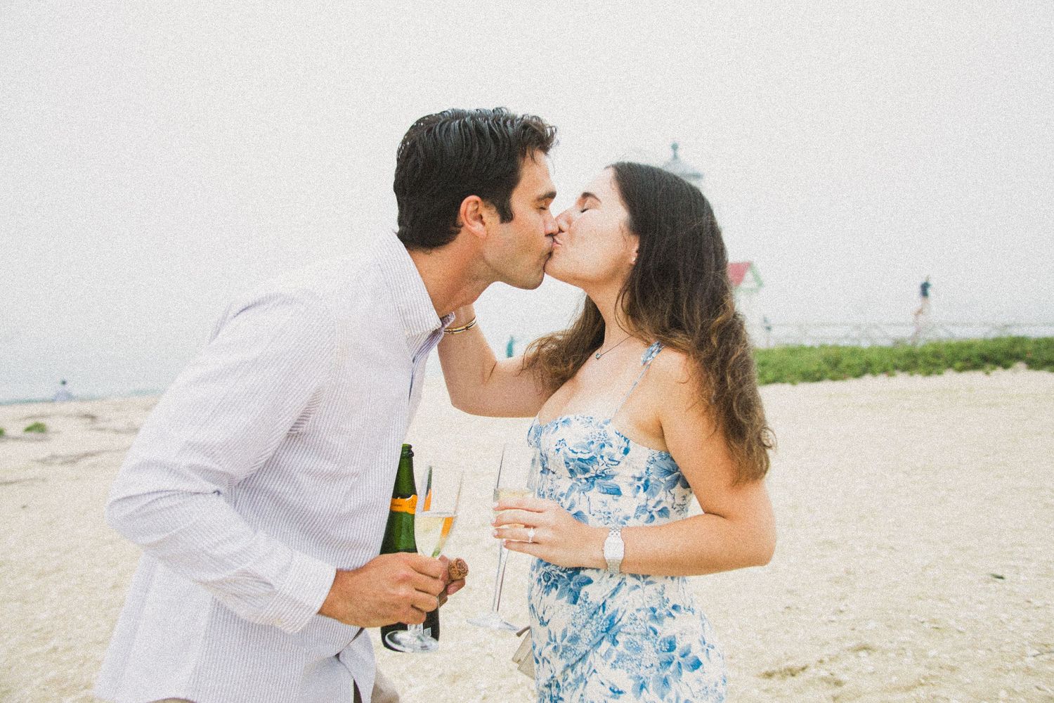 Couple kissing on beach while holding champagne glasses and wine bottle, celebrating outdoors.