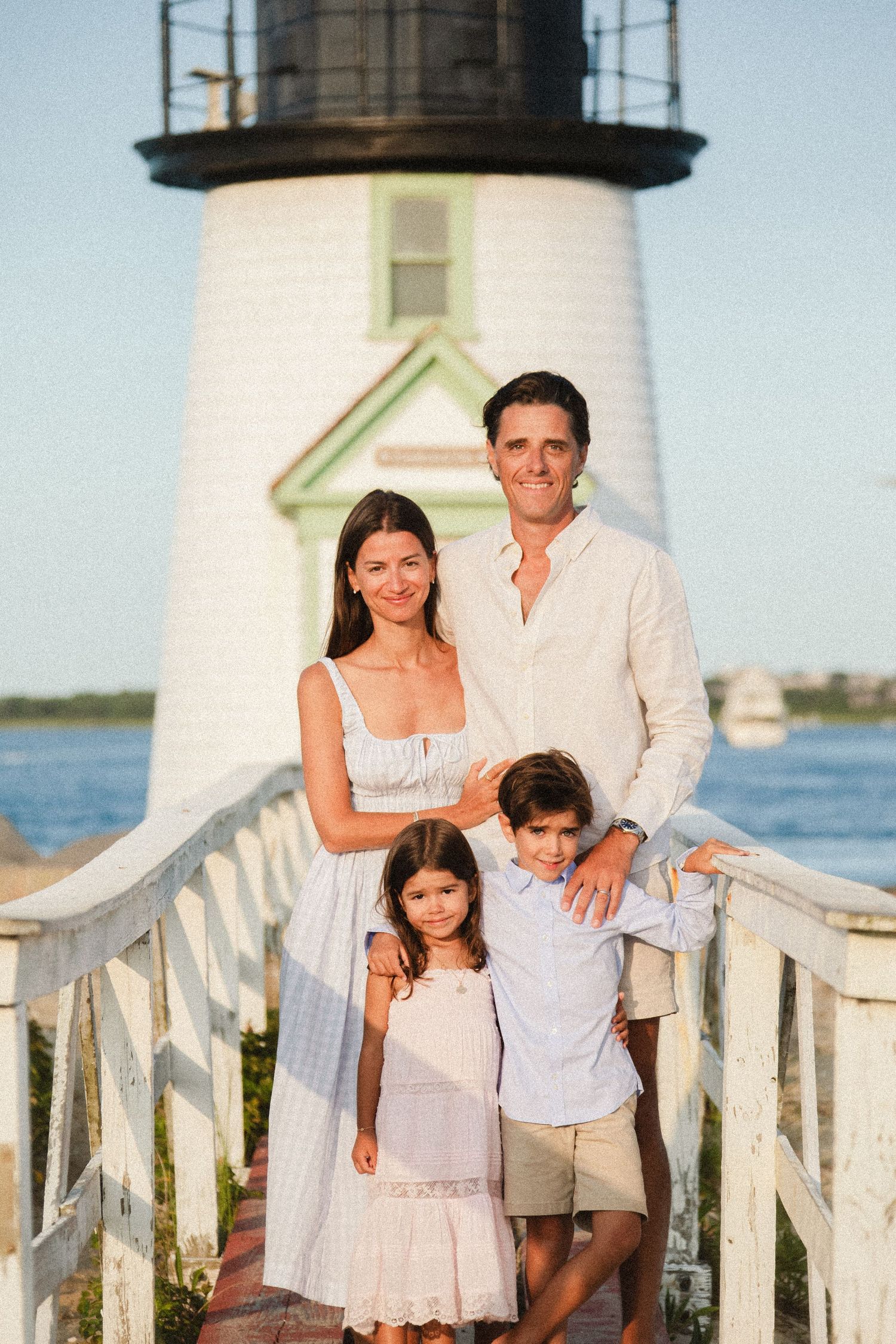 Family of four posing in front of a white lighthouse near the water on a sunny day.