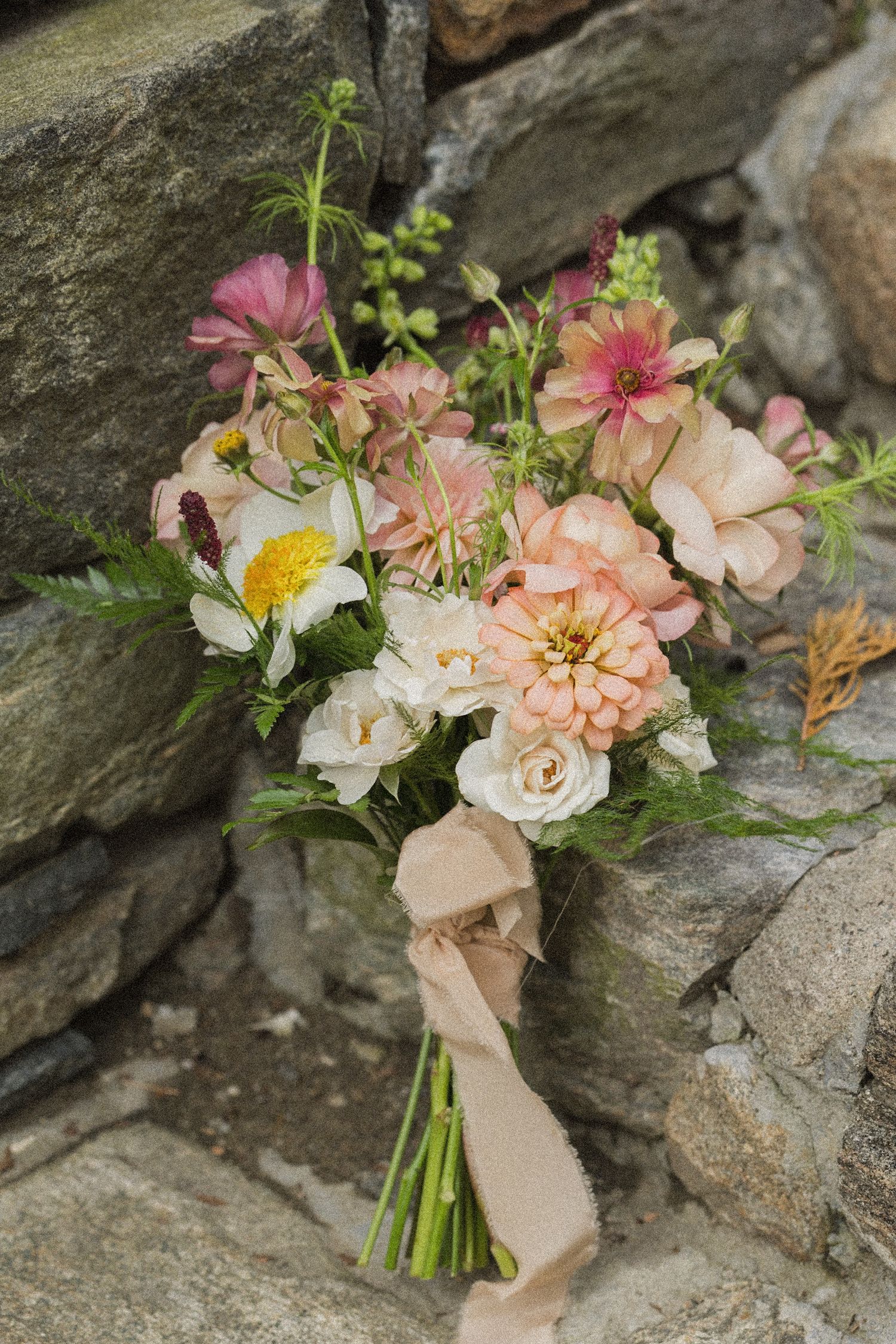 Wildflower bridal bouquet of peach dahlias, pink blooms and white daisies tied with a blush ribbon, resting against a rustic stone wall.