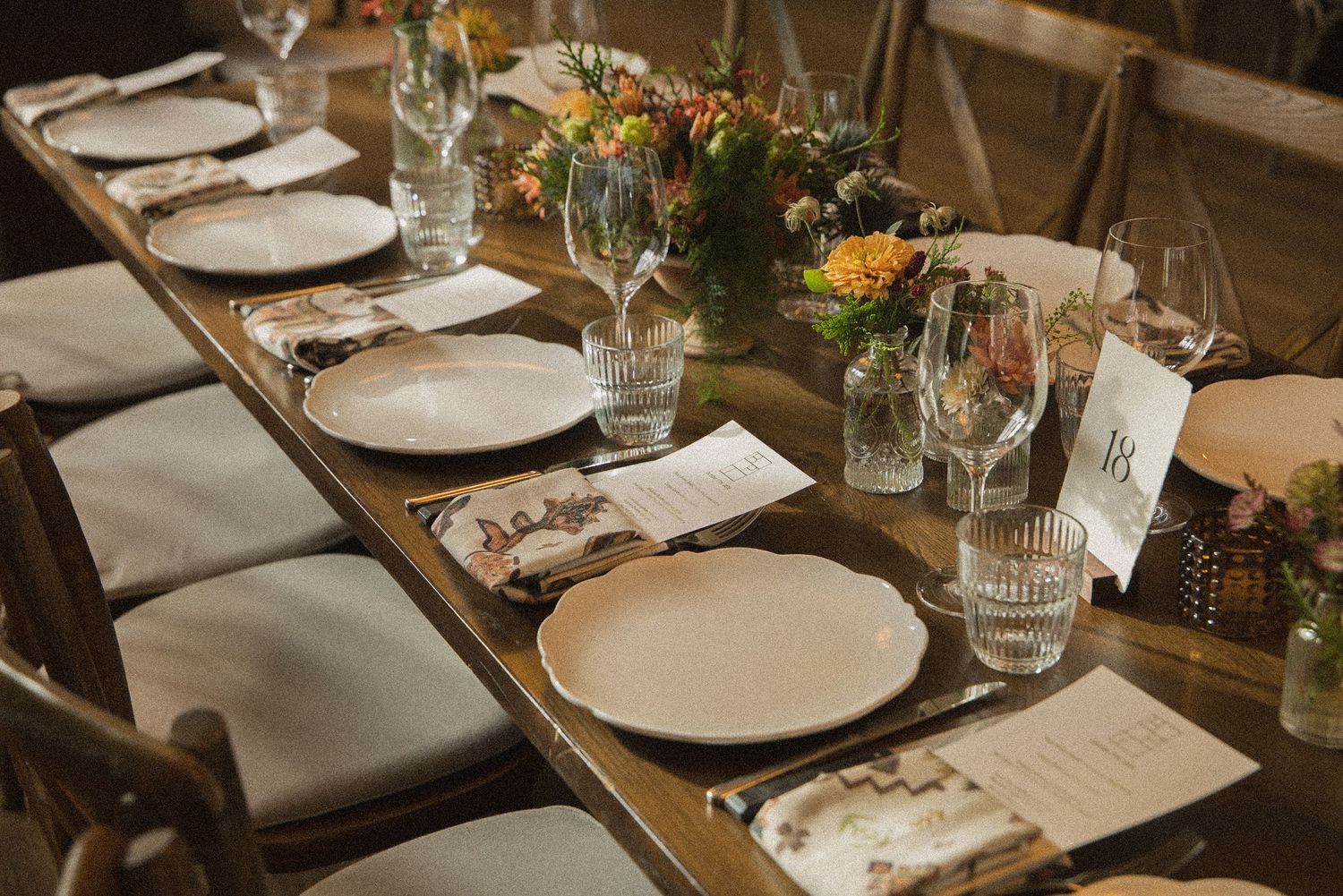 Rustic wedding reception table set with white plates, folded napkins, menus, glassware and fresh wildflower arrangements.