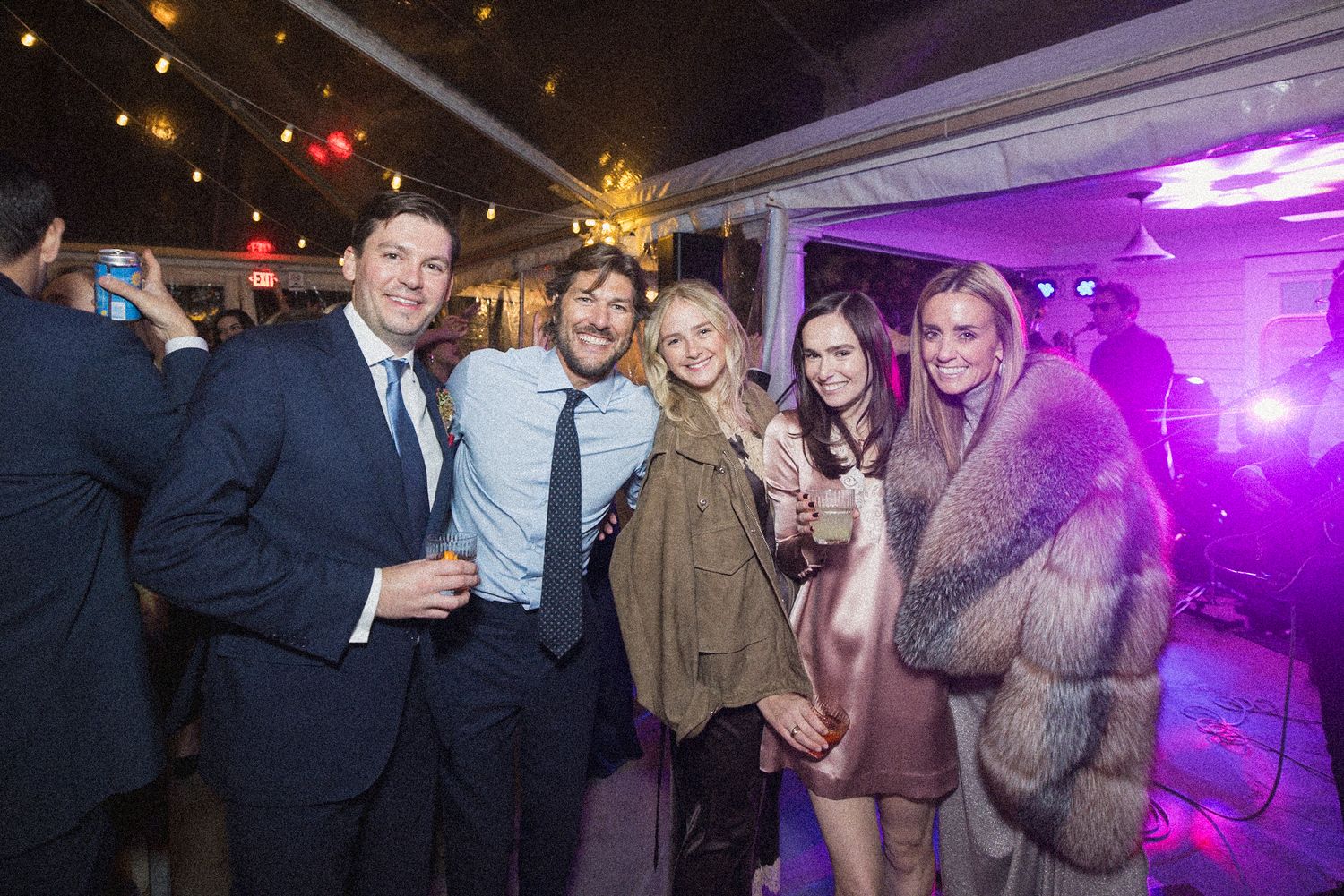 Five smiling guests in cocktail attire pose together at an evening tented event with string lights and colorful purple party lighting.