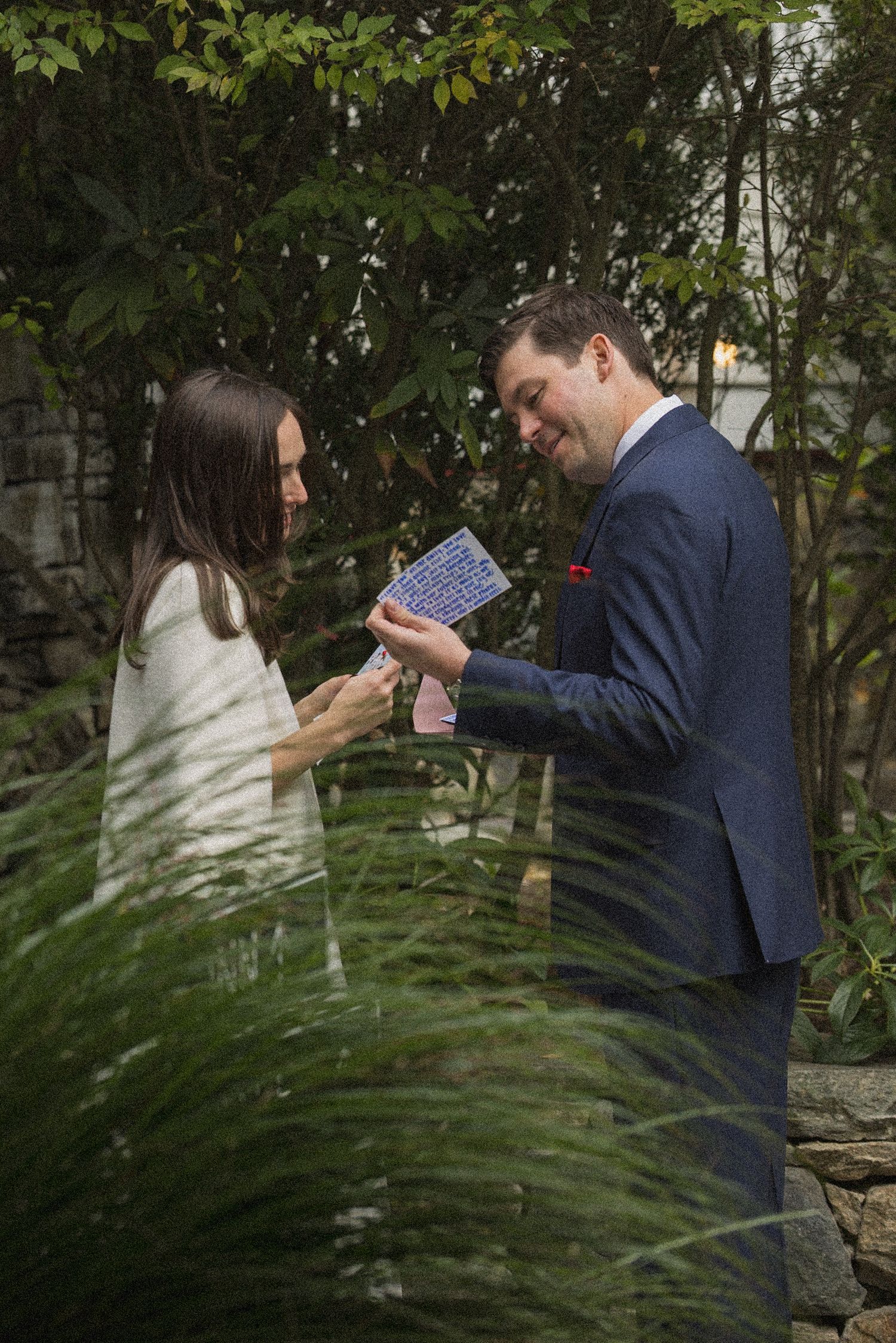 A man in a navy suit reads a handwritten card aloud to a smiling woman in white as they stand in a lush garden.