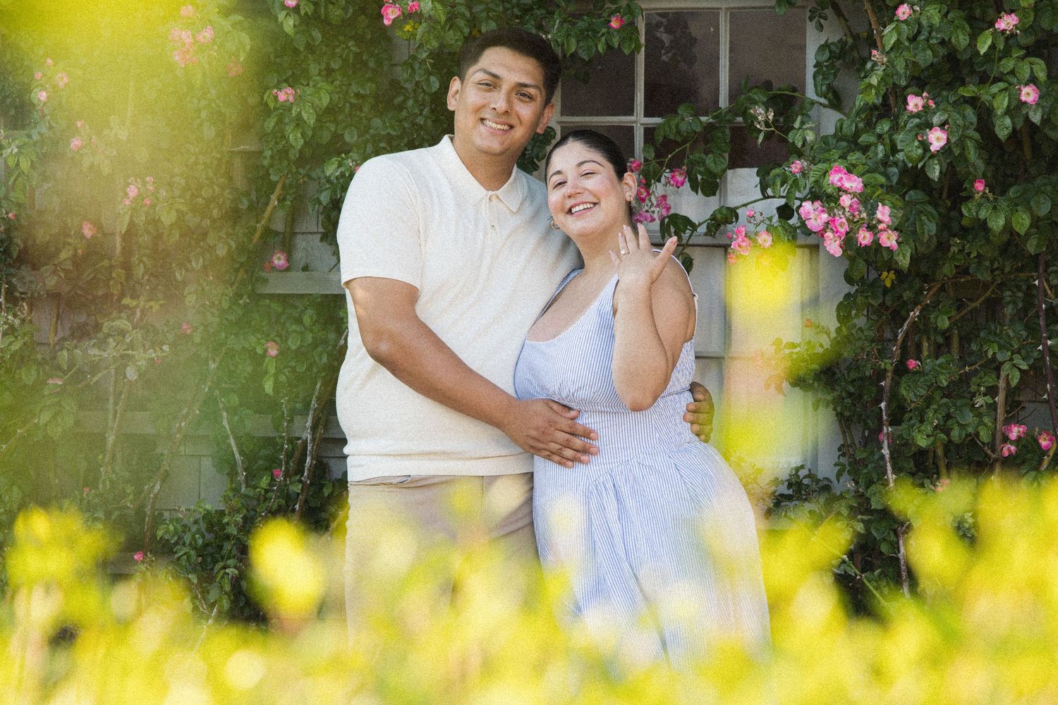A smiling couple embraces in a sunlit garden surrounded by pink roses and yellow flowers.