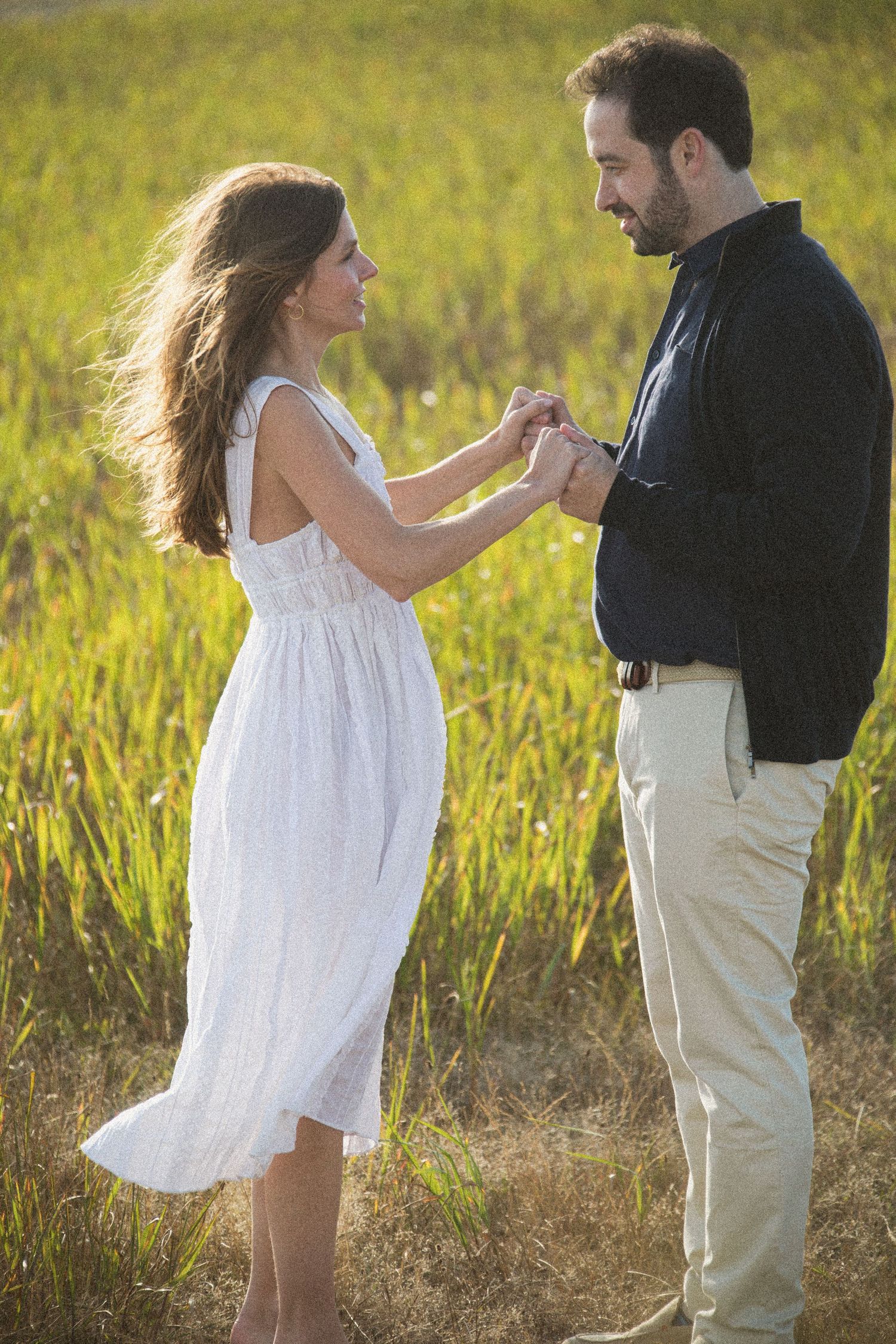 A couple dances playfully in a sunlit meadow near a coastal lighthouse.