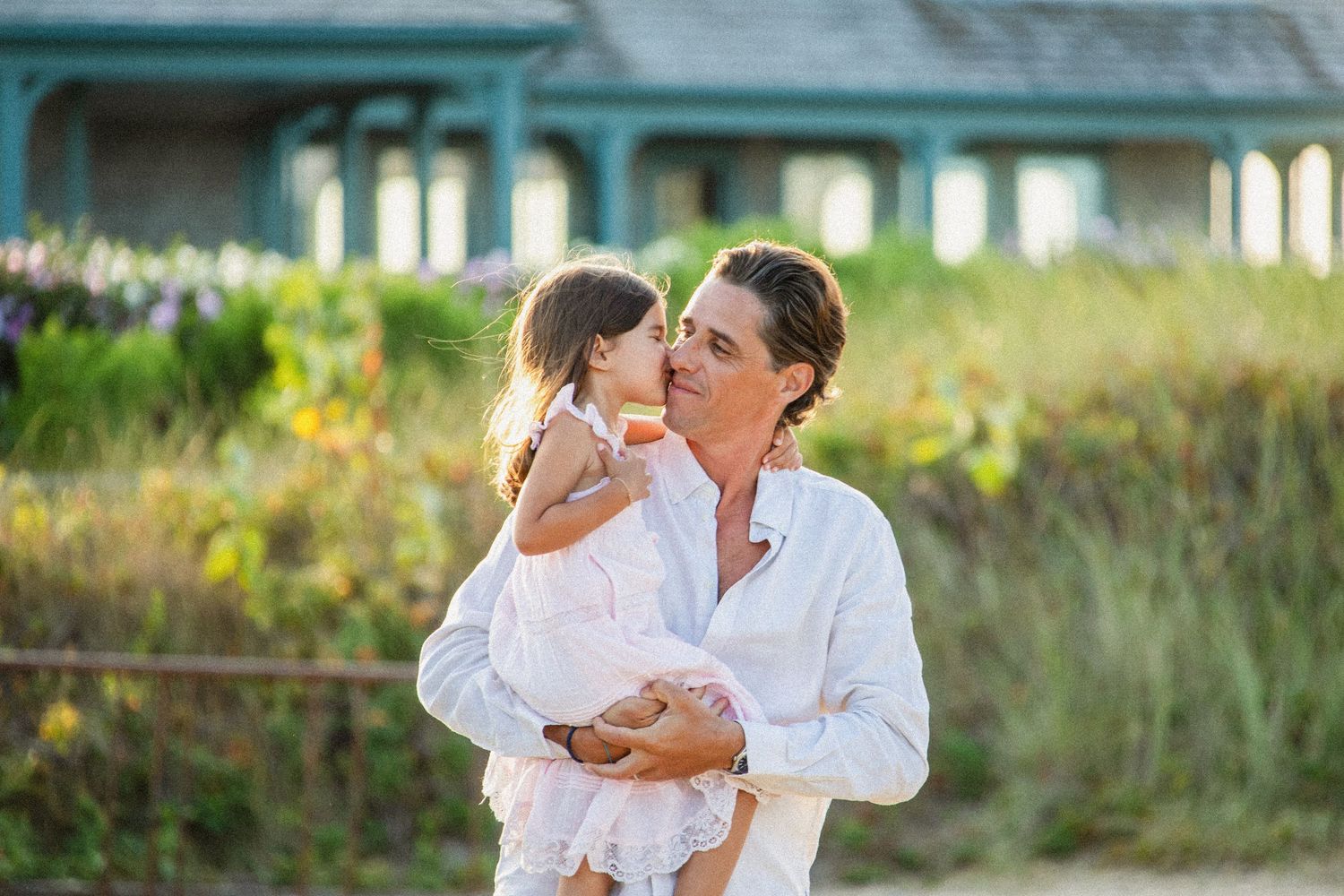 A woman in white holds a toddler girl who kisses her cheek outdoors near a green building.