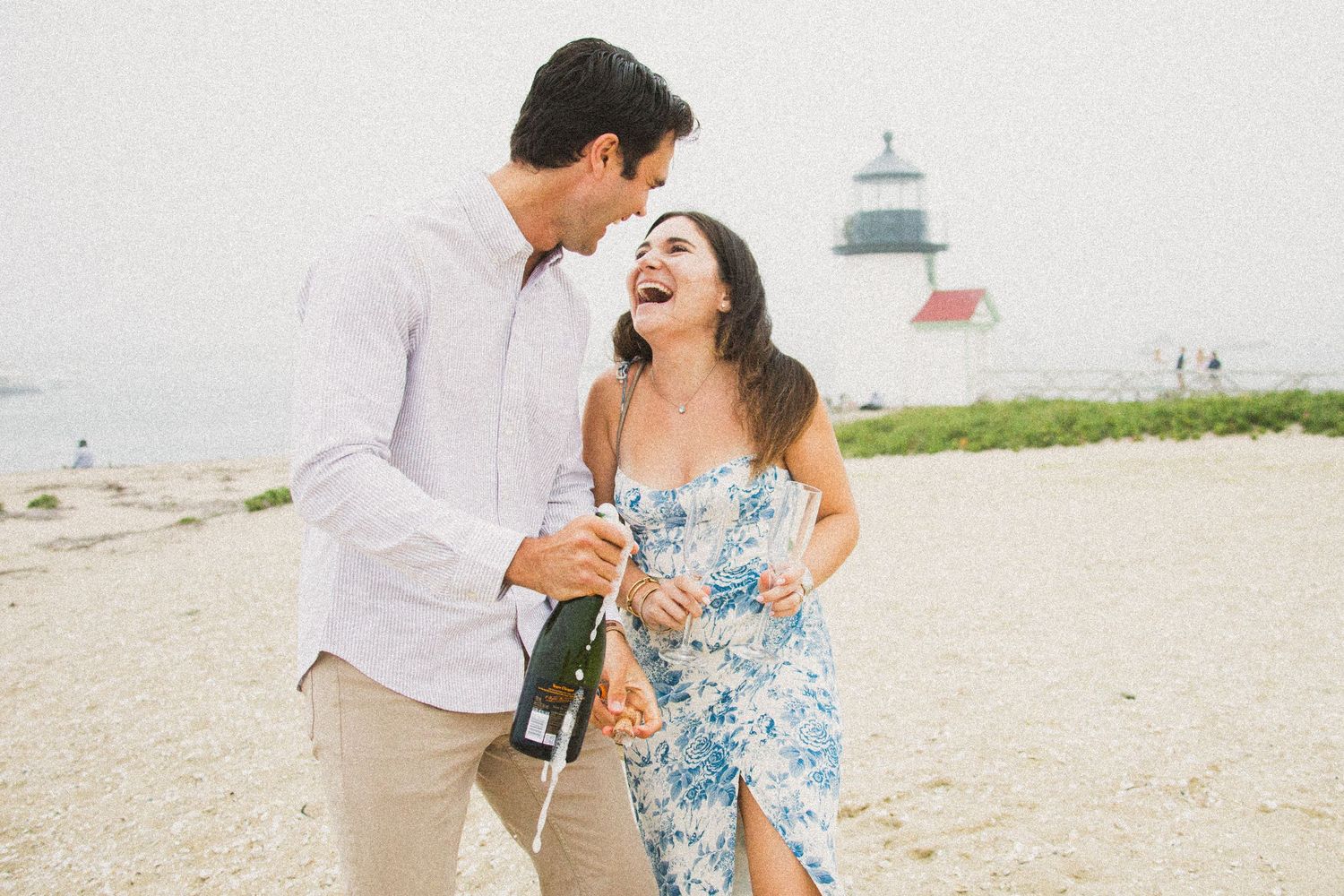 Couple laughing on beach with champagne bottle near lighthouse on overcast day.