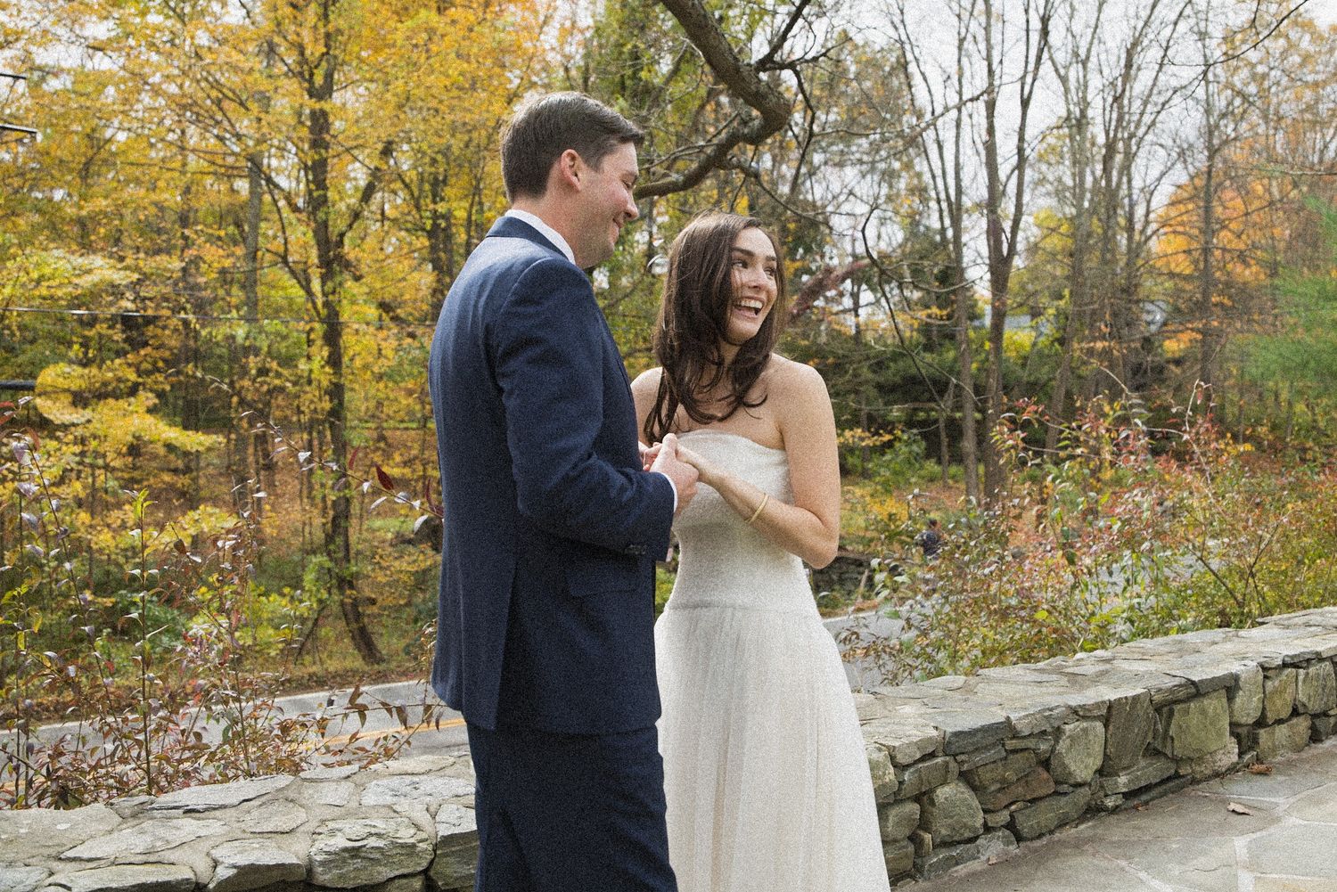 Smiling bride in a white dress and groom in a navy suit share a joyful moment beside a stone wall in a colorful autumn forest.
