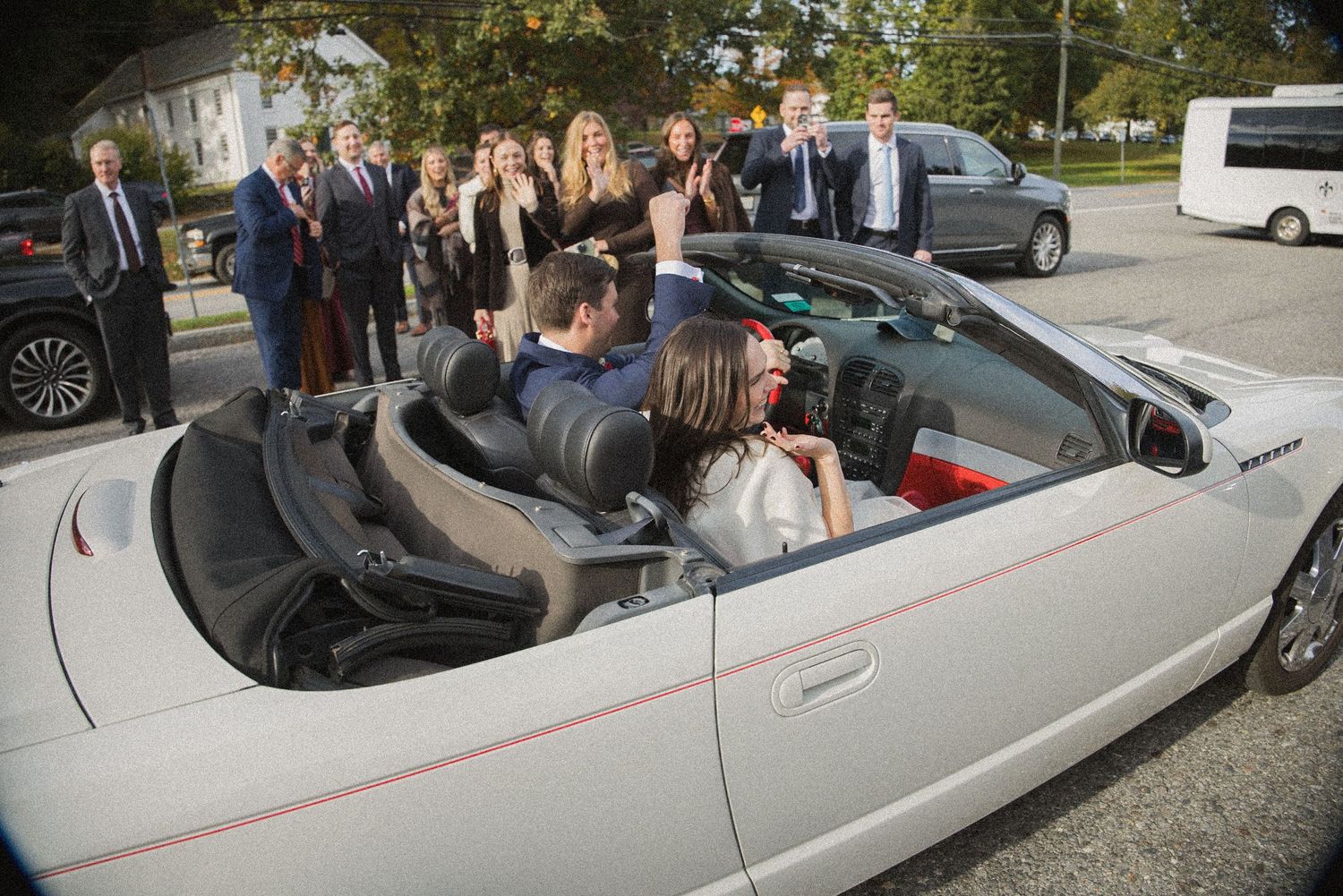 Wedding guests in formal attire gather around a white convertible as a couple sits inside during the celebration.