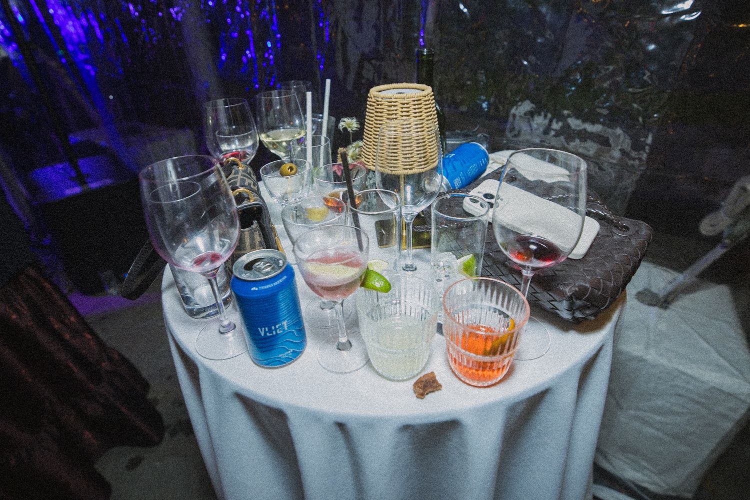 Round table with a white cloth holding half-empty wine glasses, a blue can with a straw, an orange cocktail and a wicker lid after an event.