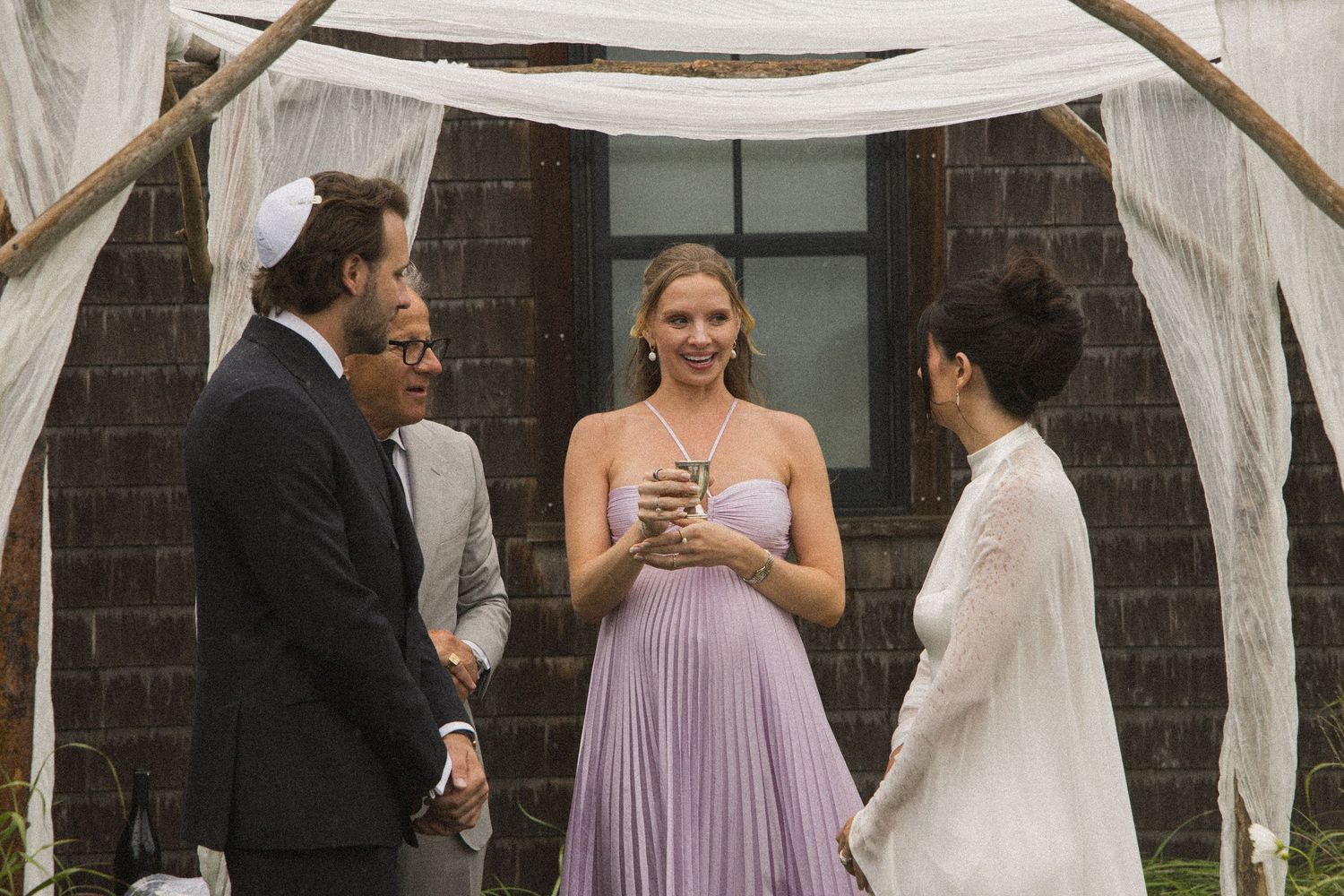 Wedding ceremony outdoors under rustic arch, woman in lavender dress holding glass, guests in formal attire smiling.