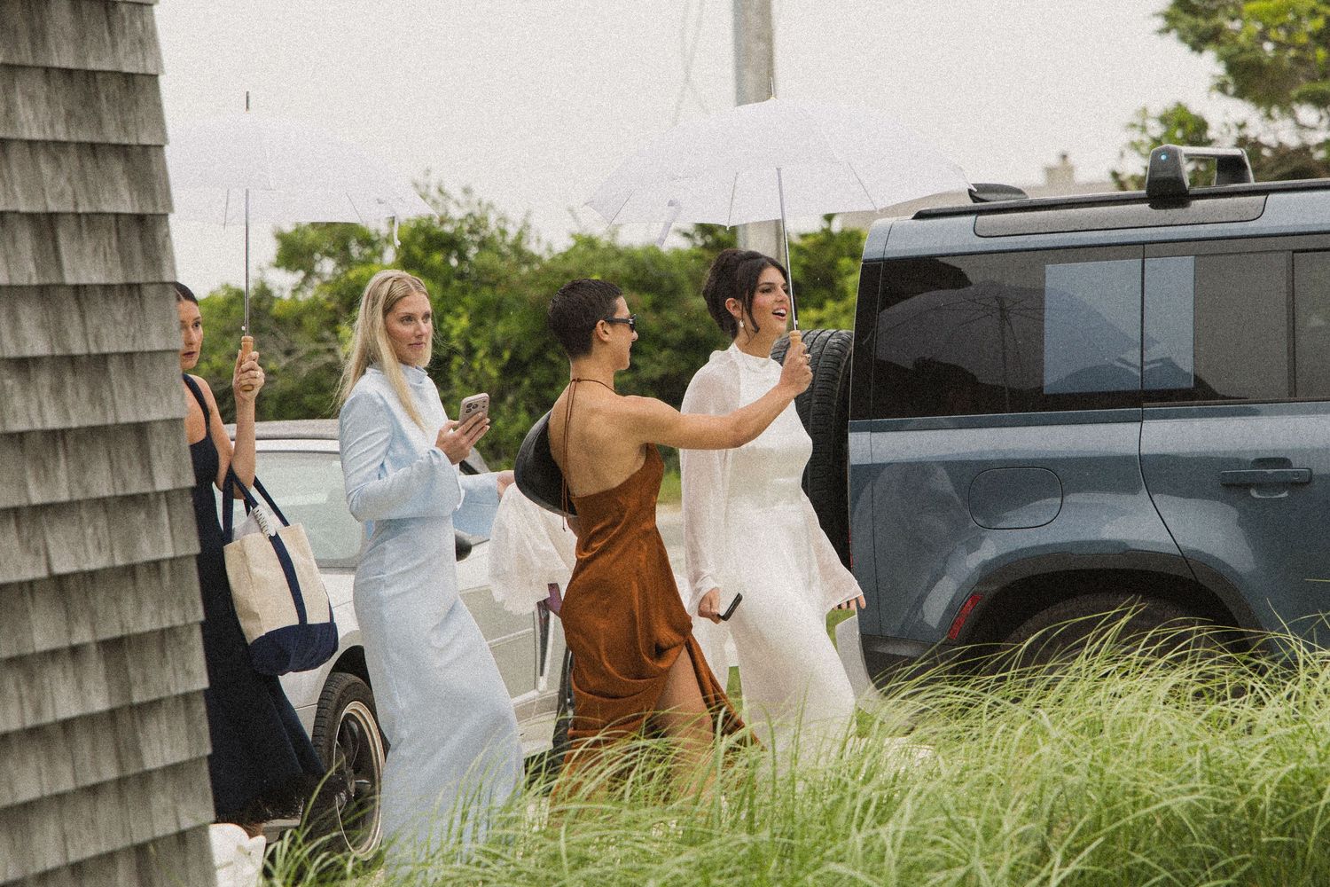 Women in elegant attire walk outdoors near a Land Rover SUV on a cloudy day, one holding a white umbrella.