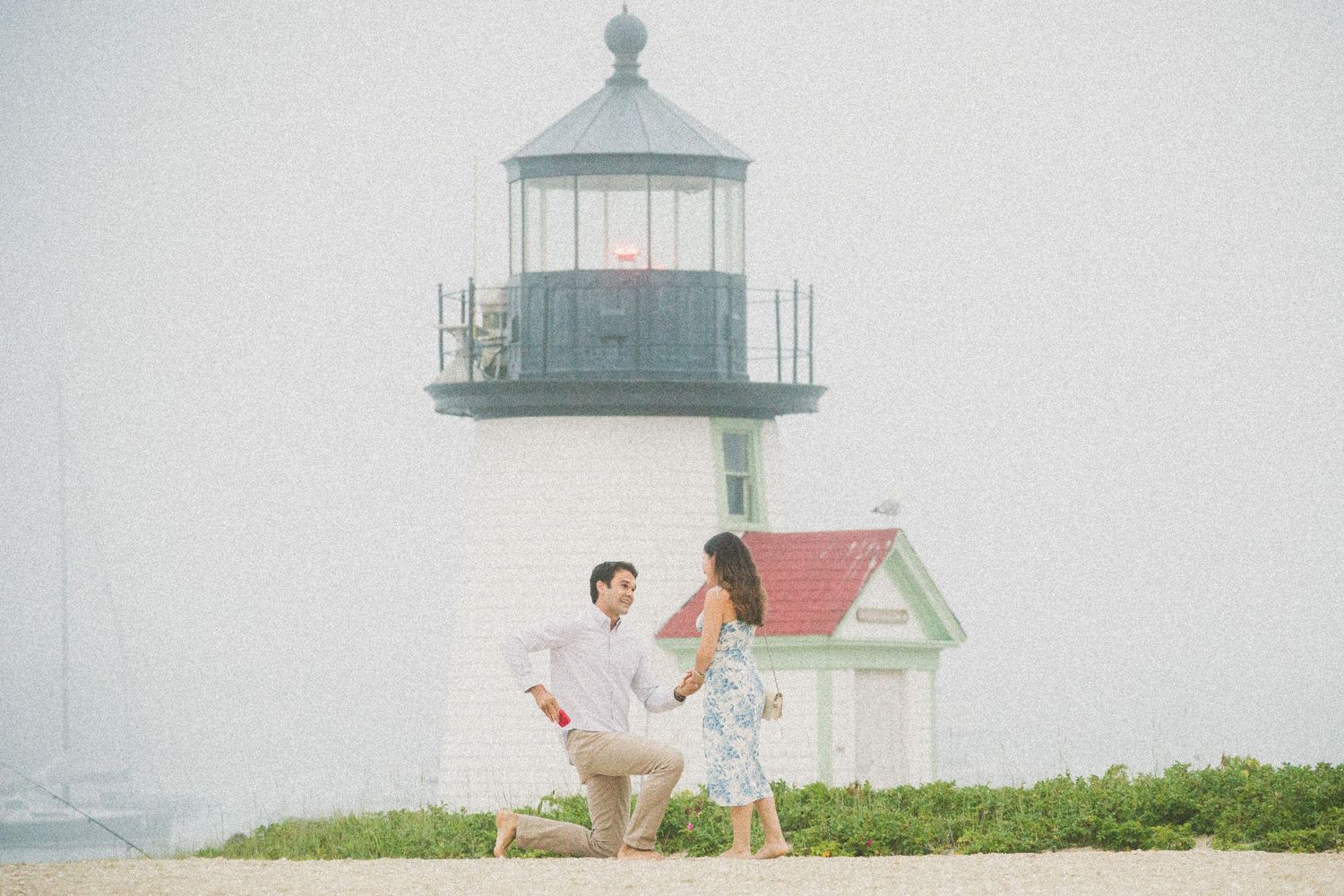 Man proposing to woman on sandy beach with white lighthouse and red-roofed building in misty background.
