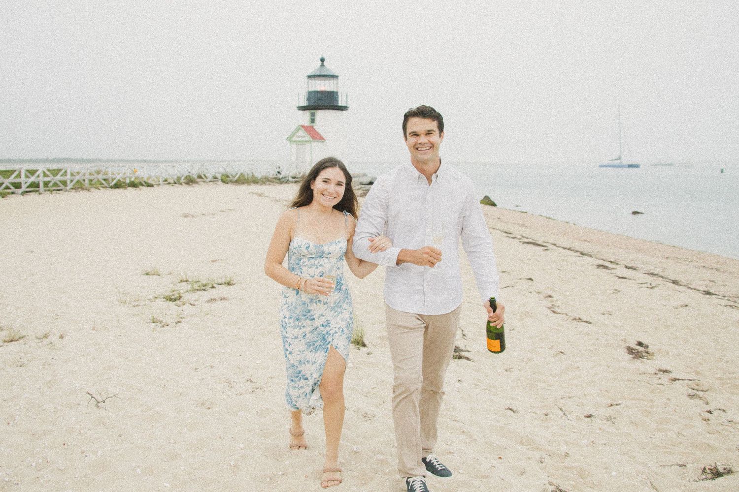 Happy couple walking on sandy beach near lighthouse, woman in floral dress, man holding champagne bottle.