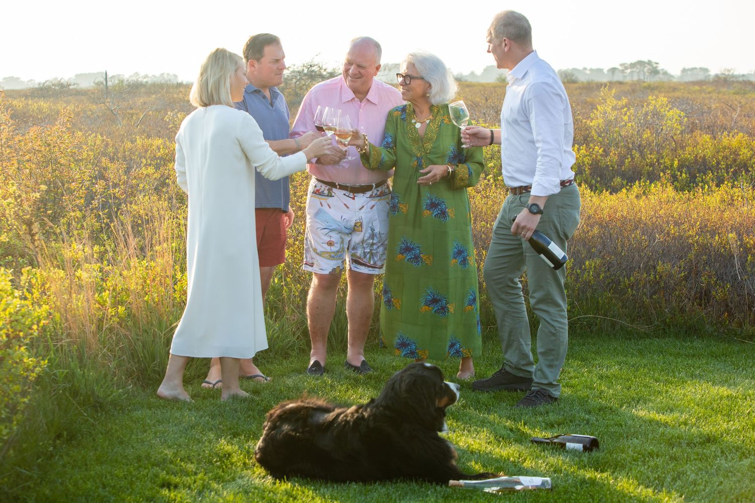 Five adults enjoying outdoor drinks on green grass at sunset with a black dog resting nearby amid golden foliage.
