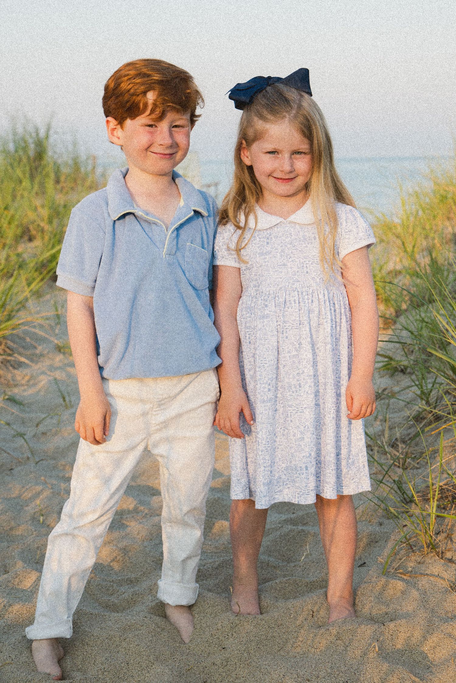 Two children, a red-haired boy in blue polo and a blonde girl in floral dress, pose together on a sandy beach.