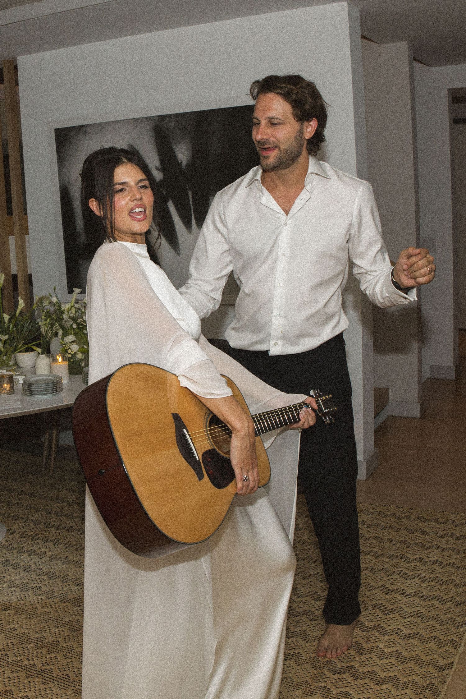 Couple dancing together at indoor event, woman in white dress holding acoustic guitar, man in white shirt and black pants.