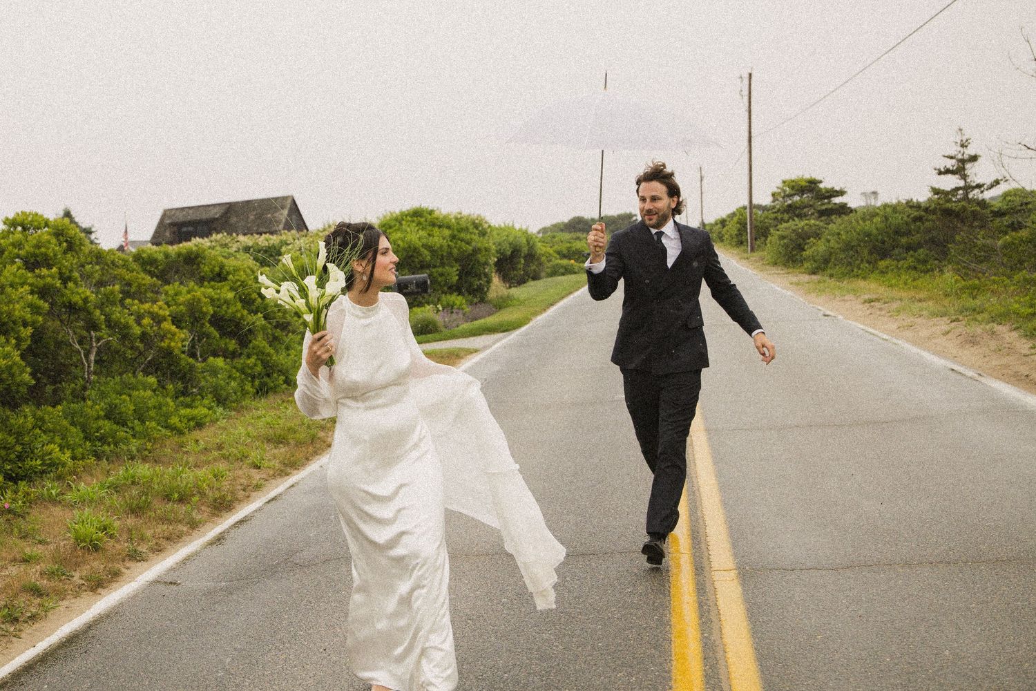 Bride and groom walking on a rainy road, bride in white gown holding flowers, groom in black suit with umbrella.