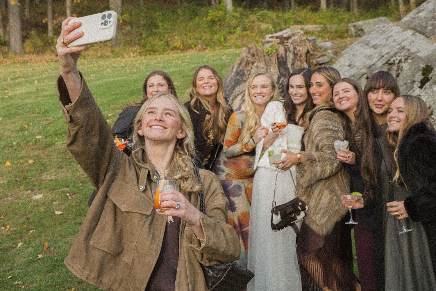 A smiling woman in an olive jacket takes a group selfie with friends holding drinks on a sunny grassy lawn in autumn.