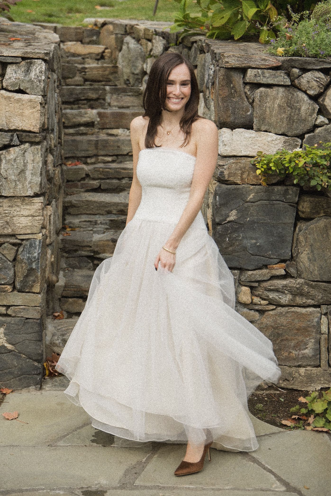 Smiling woman in a strapless white tulle gown and brown pointed flats stands on a stone patio beside a rustic stone wall and steps.