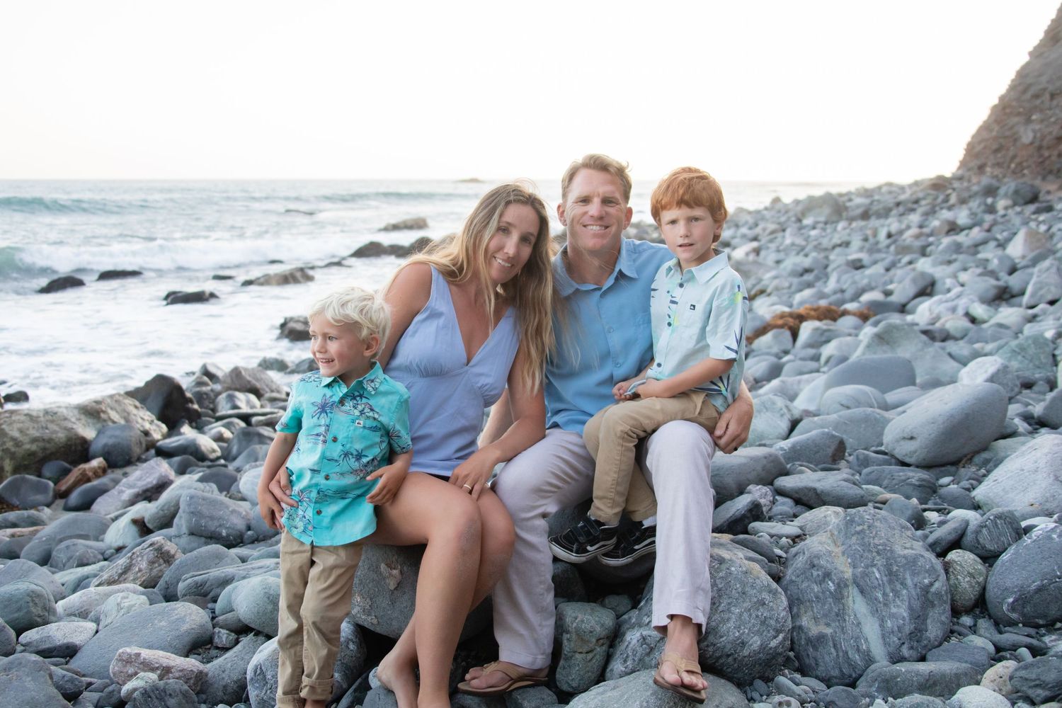 A family poses for a portrait while sitting on large rocks at a beach.