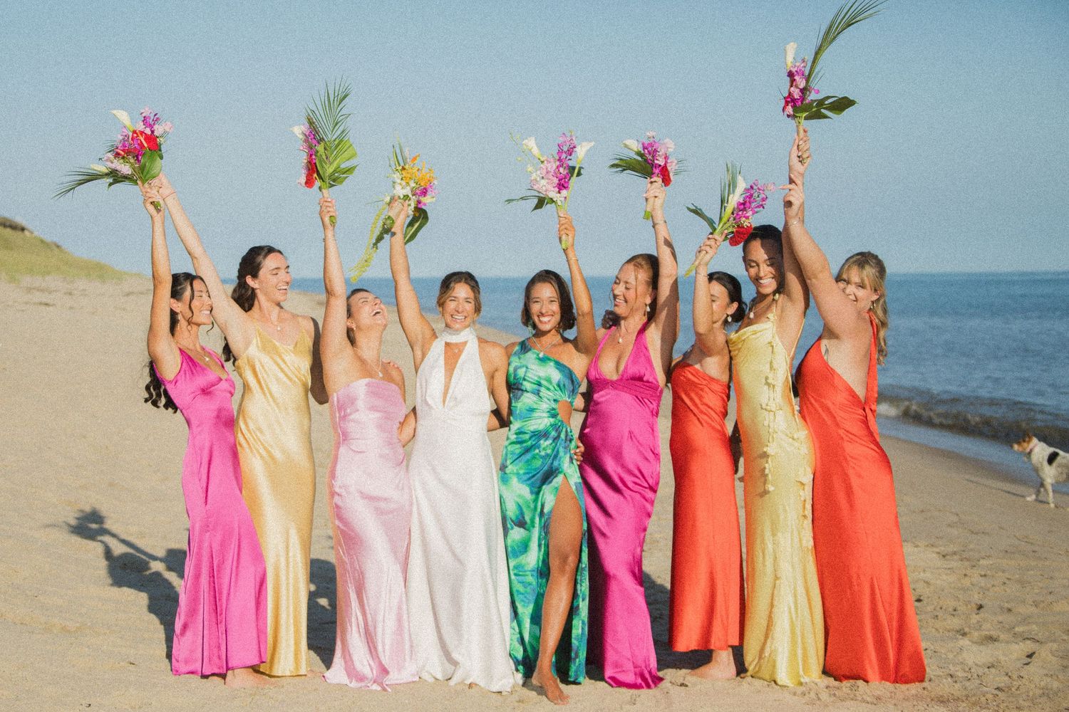 Wedding party in colorful gowns raising tropical bouquets on a sunny beach.
