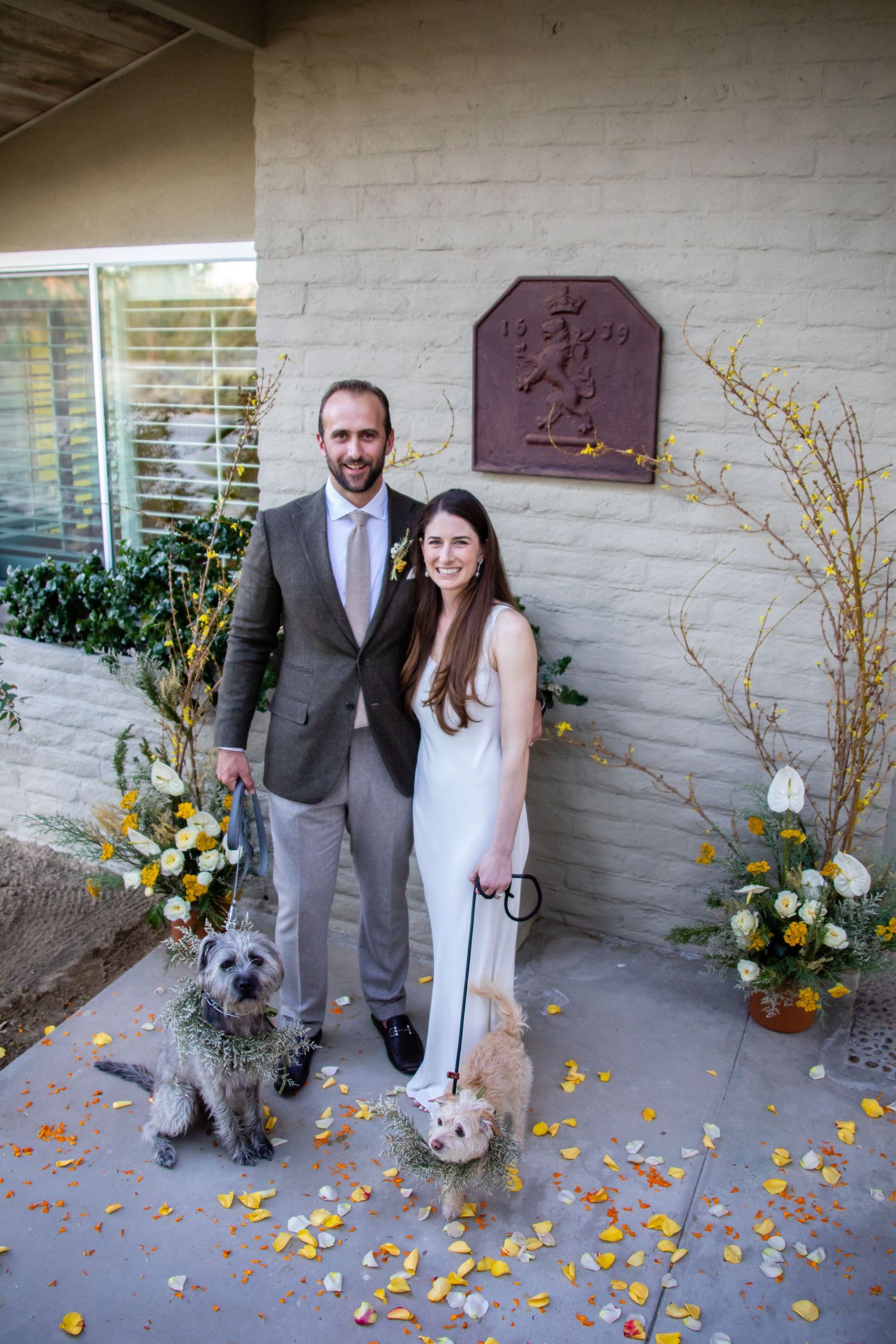 A couple and their dog stand among scattered flower petals against a light colored wall.