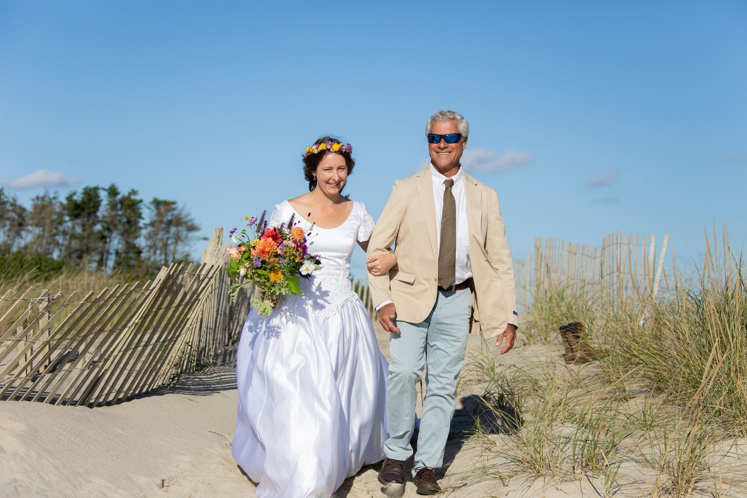 A couple walks along a sandy beach path with beach grass and blue skies in the background.