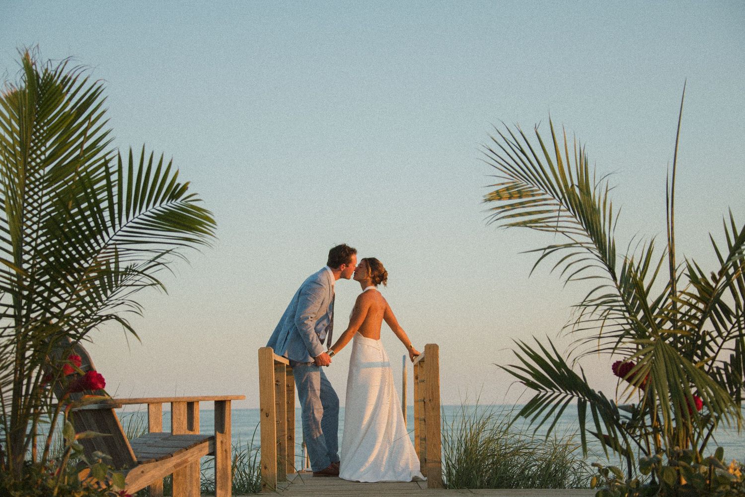 A couple stands together by wooden fence posts with palm fronds framing the coastal scene.