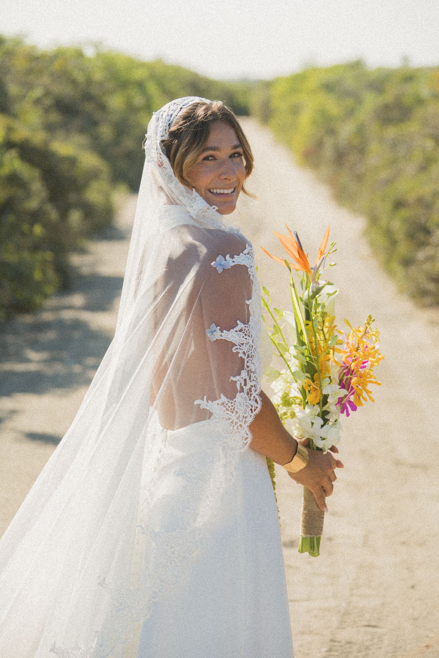 A bride in a lace-detailed wedding dress with long veil smiles while holding a vibrant tropical bouquet on a sunny path.