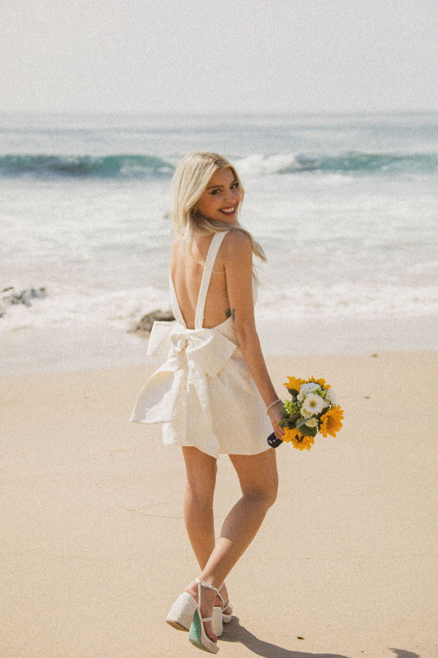 Bride in short white dress poses playfully with sunflowers on sandy beach.