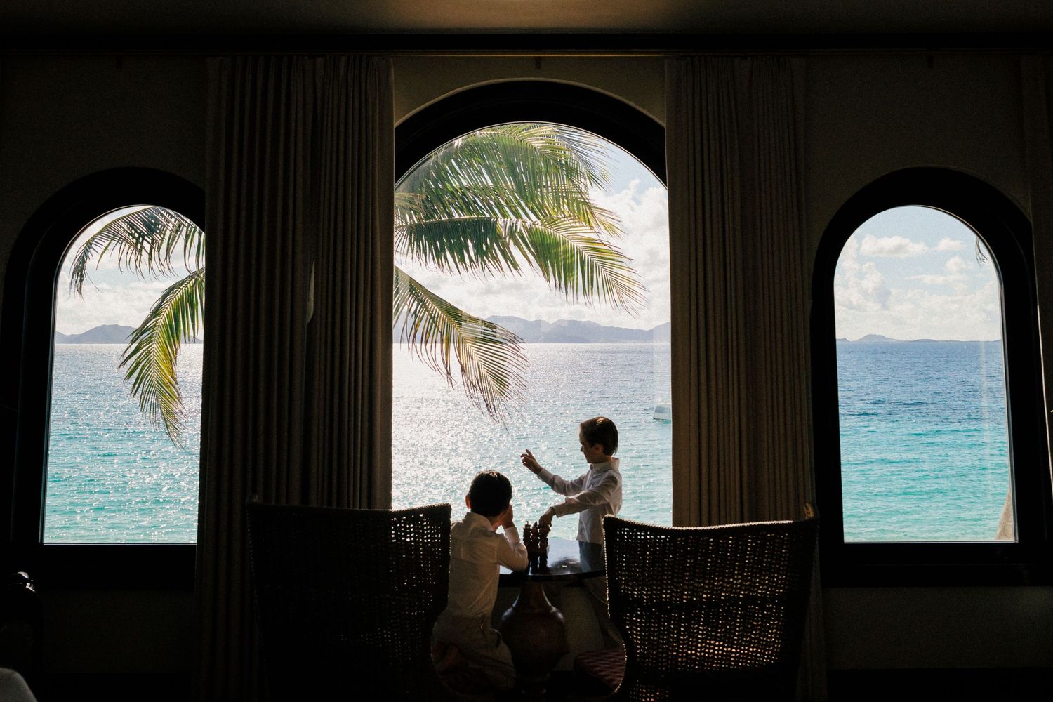 Silhouetted arched windows frame a tropical ocean view with palm trees.