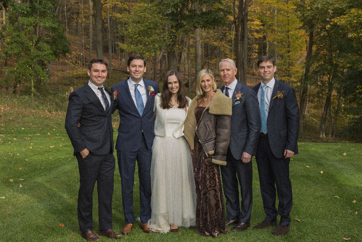 Bride in white gown poses with four men in navy suits and a blonde woman on a grassy lawn surrounded by autumn trees at an outdoor wedding.