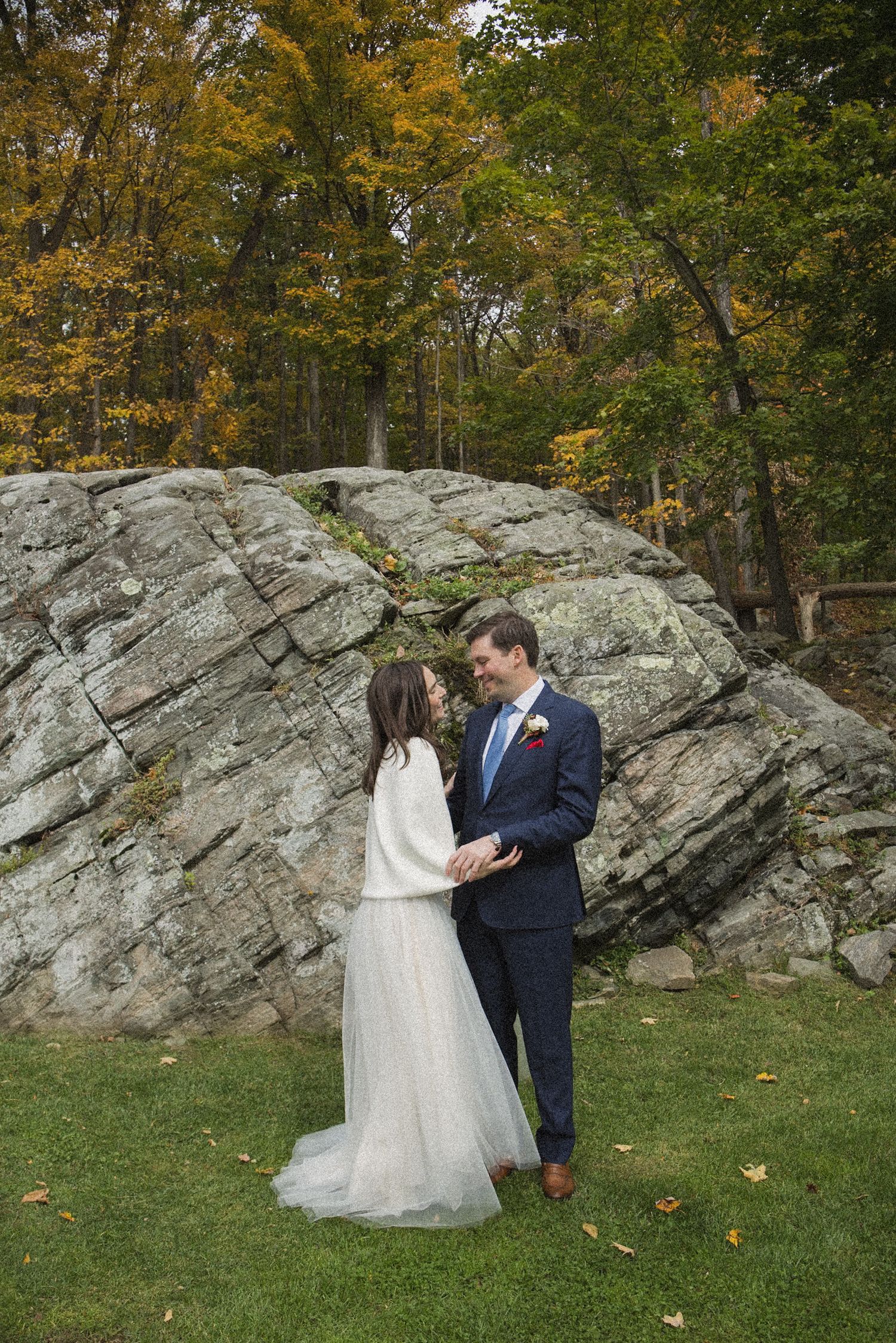 Bride in a white tulle gown and sweater holds hands with groom in a navy suit on a lawn before a large rock and autumn trees.