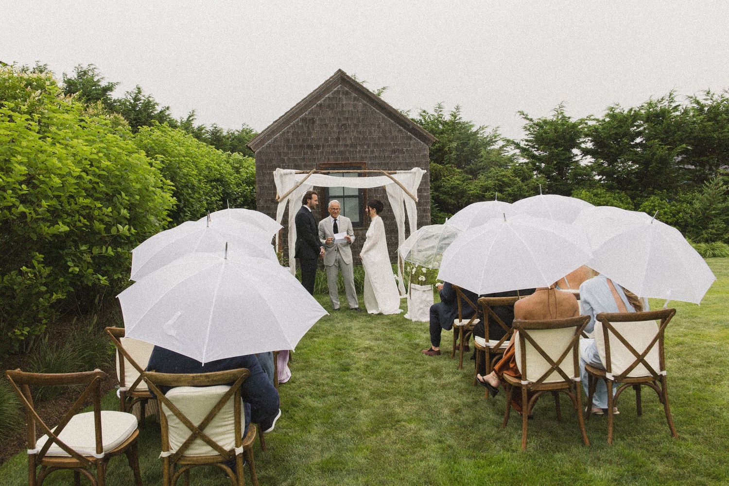 Outdoor wedding ceremony on green lawn with guests holding white umbrellas on a rainy day, rustic barn backdrop.