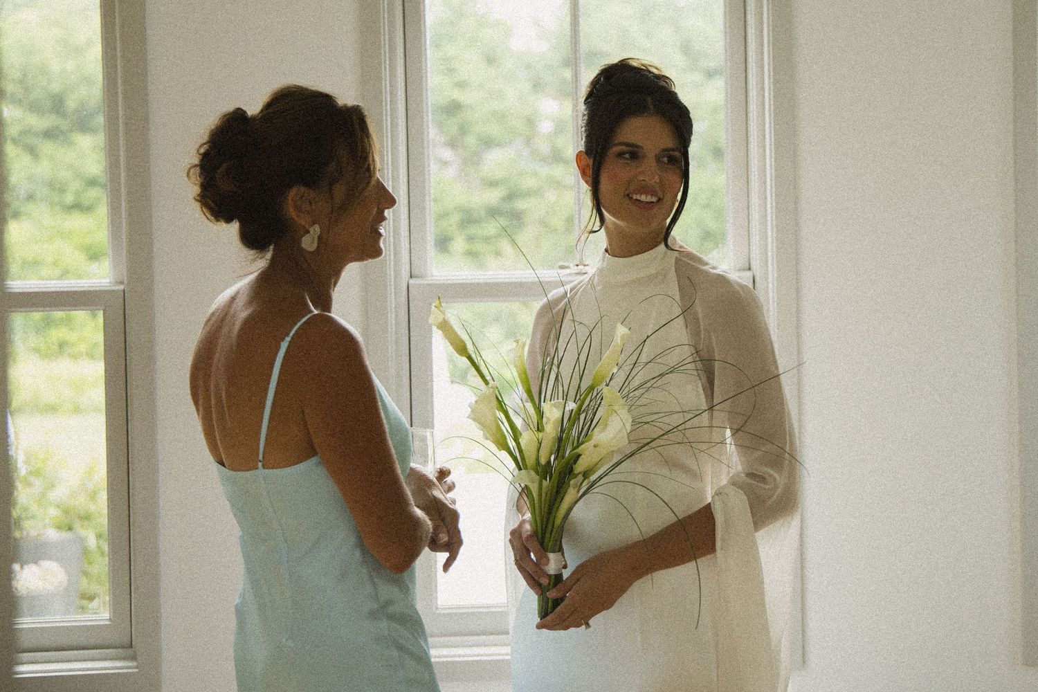 Bride in white dress holding flowers talks with bridesmaid in blue gown near bright windows.