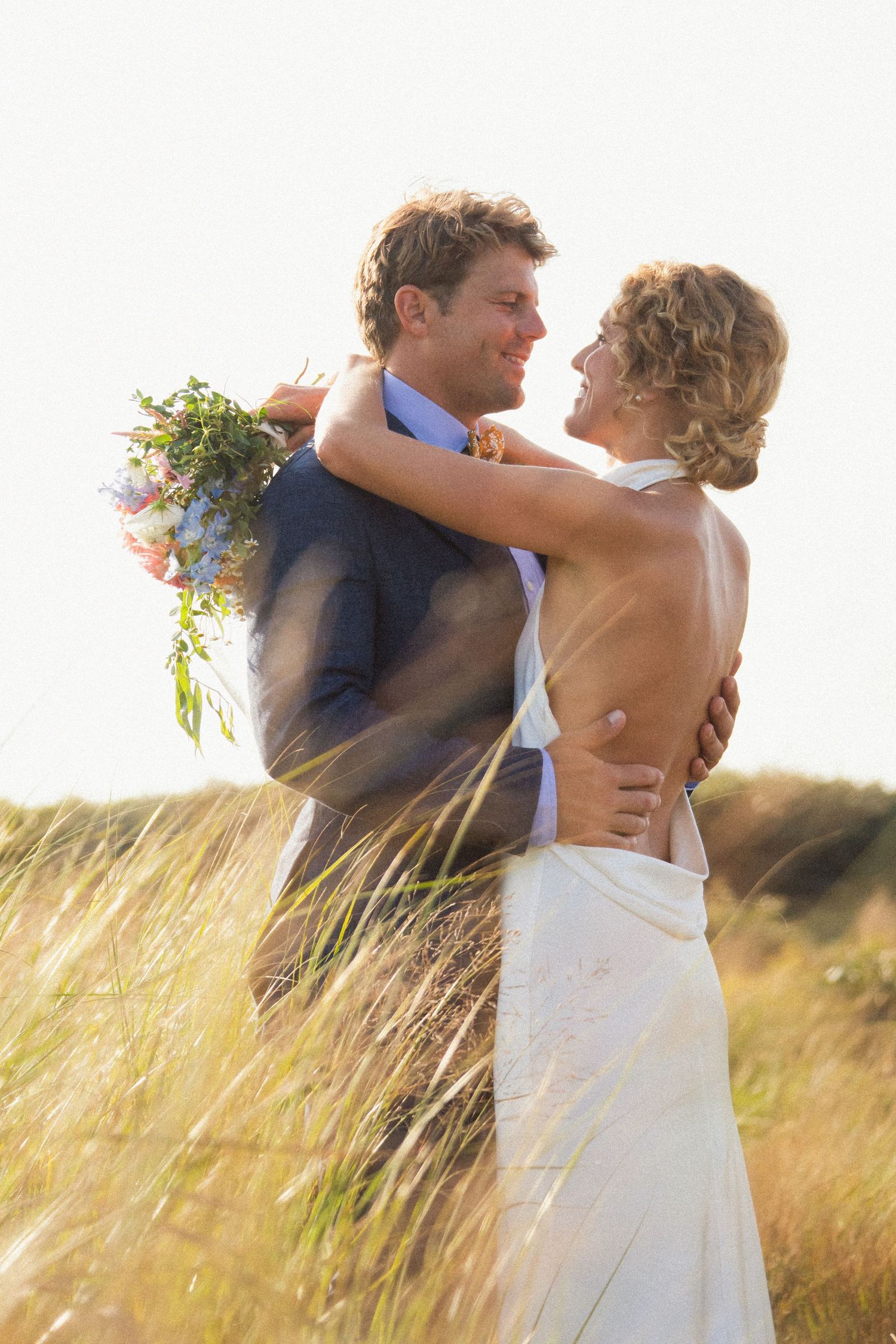 A bride and groom embrace romantically in a golden field, with the bride holding a colorful bouquet on a sunny day.