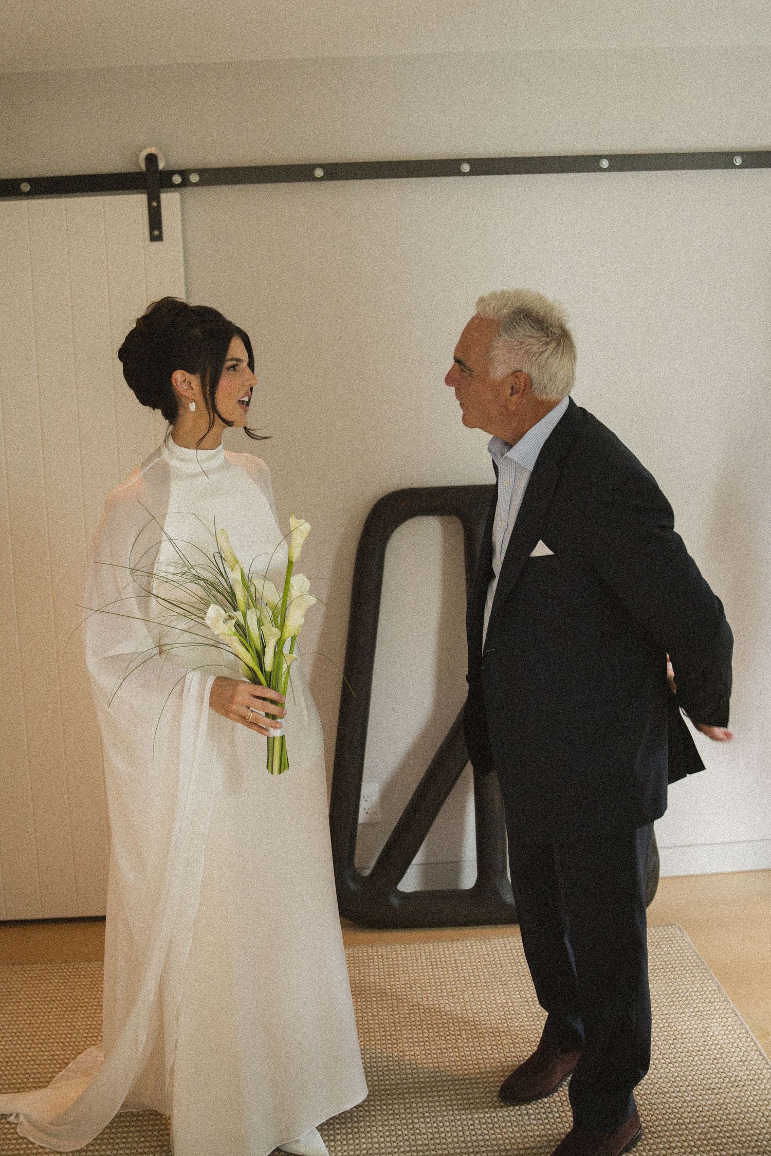 Bride in white gown holding calla lilies bouquet shares a joyful moment with an older gentleman in a dark suit.