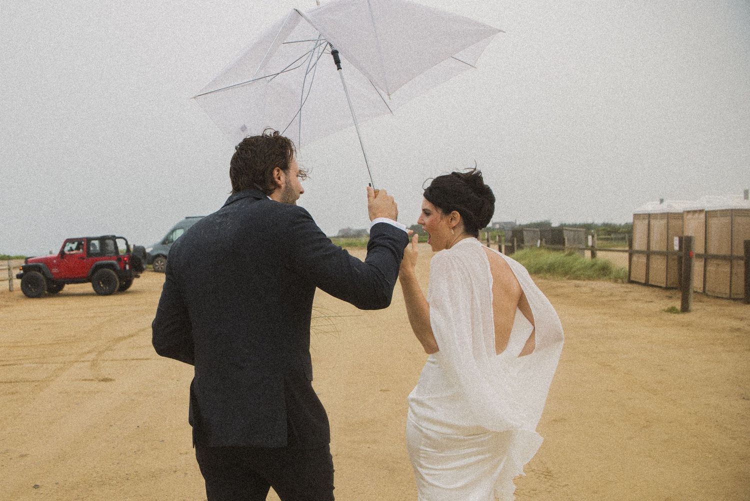 A bride and groom walk together under a white umbrella on a sandy beach on a rainy, overcast wedding day.