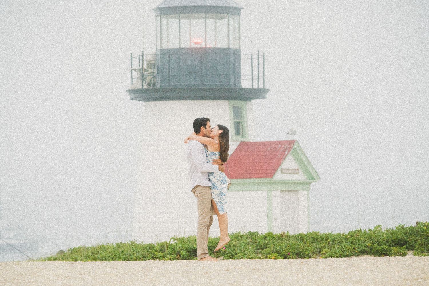 Couple sharing a romantic kiss in front of a historic lighthouse on a foggy beach day.