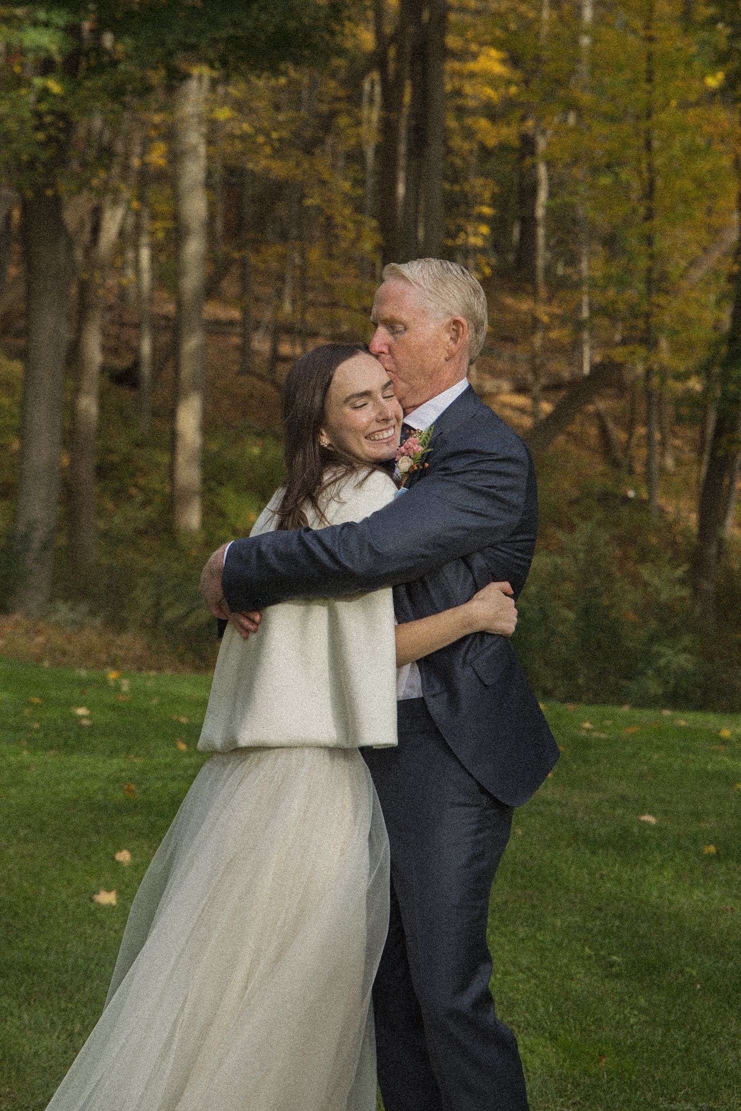 Bride in a white tulle skirt and shawl embraces an older man in a navy suit as he kisses her forehead on a lawn near autumn trees.